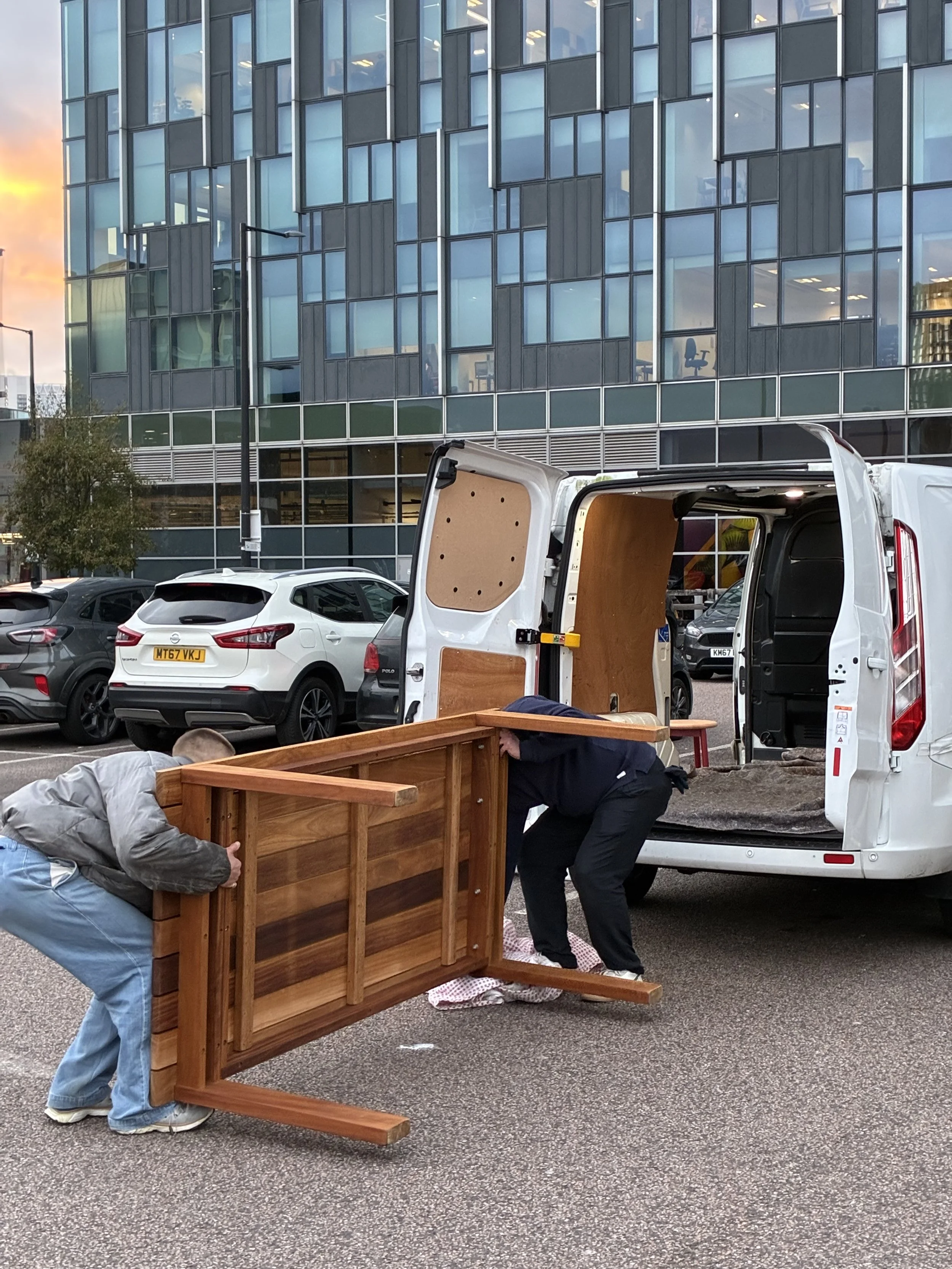 People are moving a large wooden furniture piece into the back of a white delivery van in a parking lot, with a modern glass building in the background.