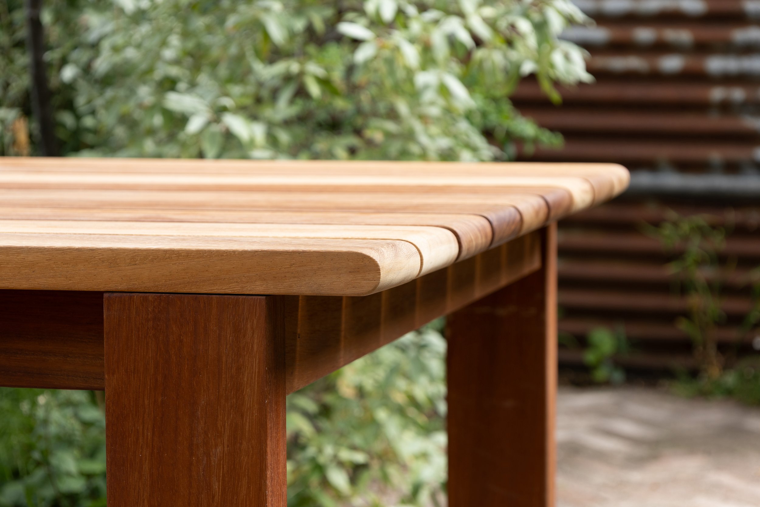 Close-up of a wooden outdoor furniture table with a background of green foliage and a wooden fence.