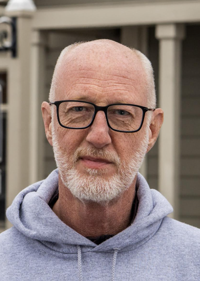 Close-up of an older man with glasses and a white beard, wearing a gray hoodie, standing outdoors in front of a house with beige siding.