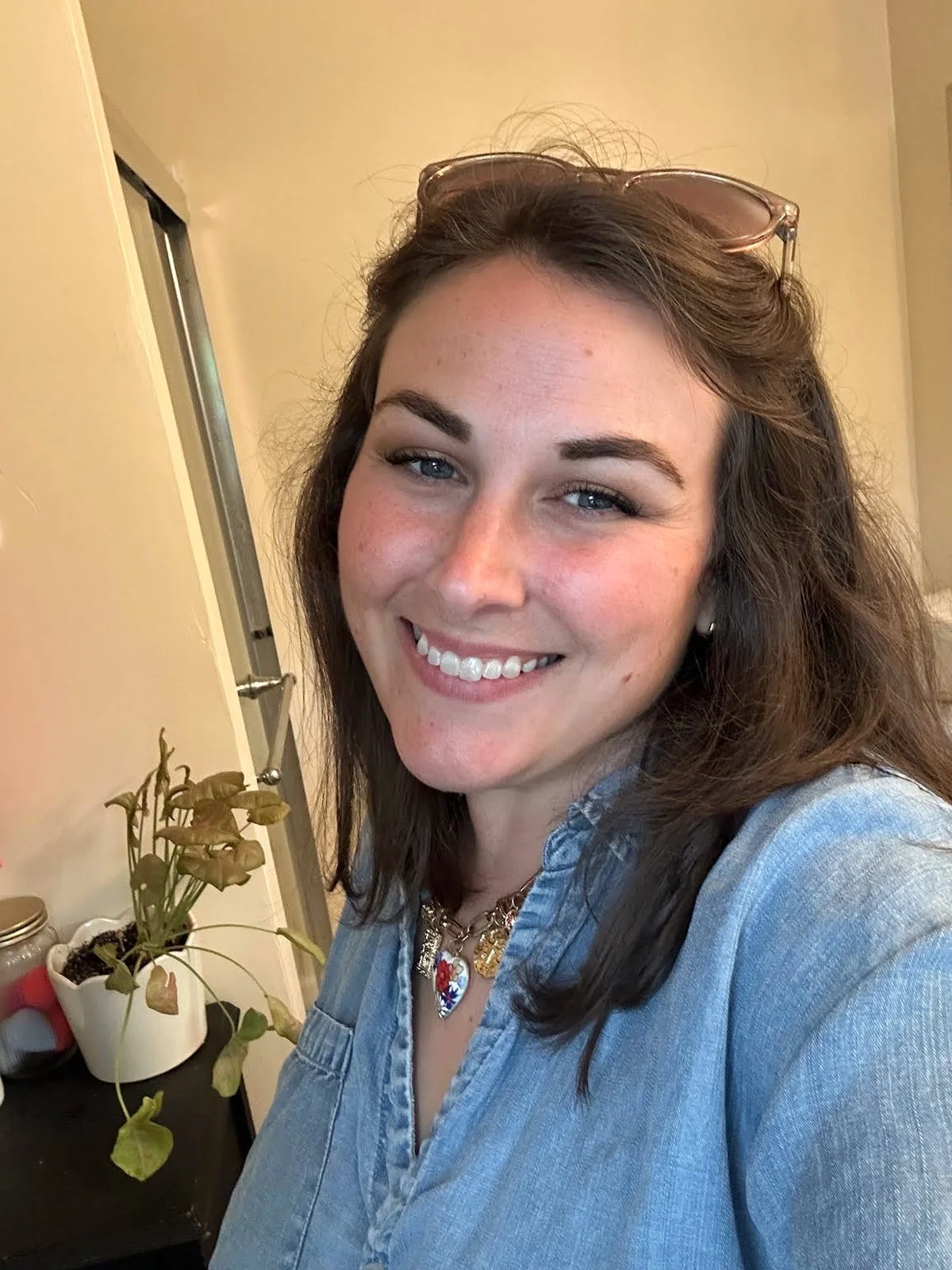 A young woman smiling with sunglasses on her head, wearing a denim shirt and a colorful necklace. Behind her is a yellow wall and a small table with potted plants.