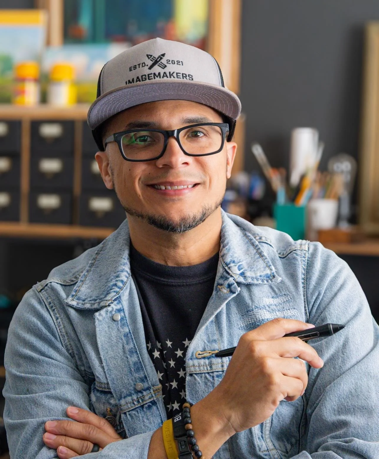 A smiling man with glasses wearing a gray baseball cap and denim jacket, holding a pen in a workspace with shelves and art supplies in the background.