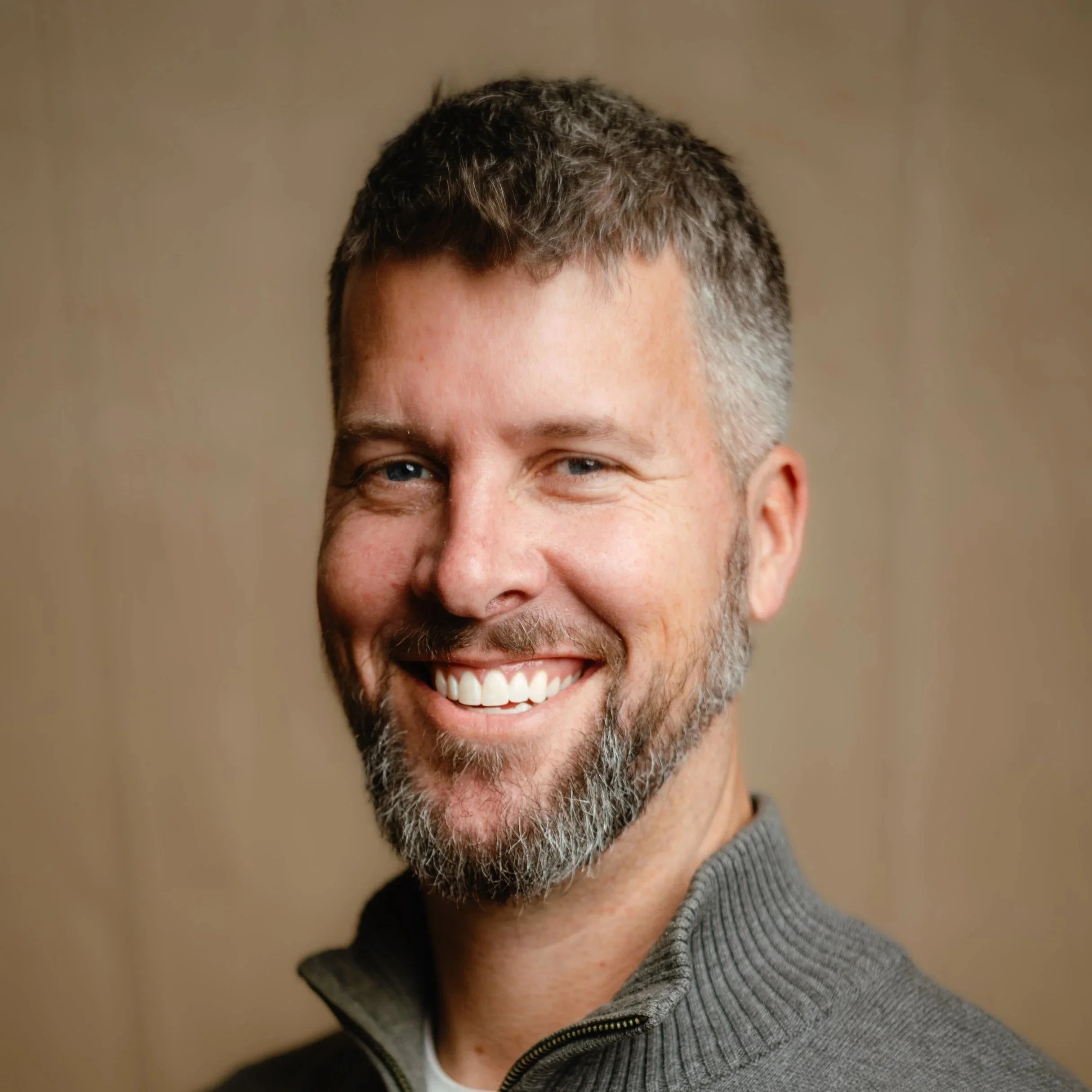 Close-up of smiling man with short, dark, and gray hair, wearing a gray hoodie.