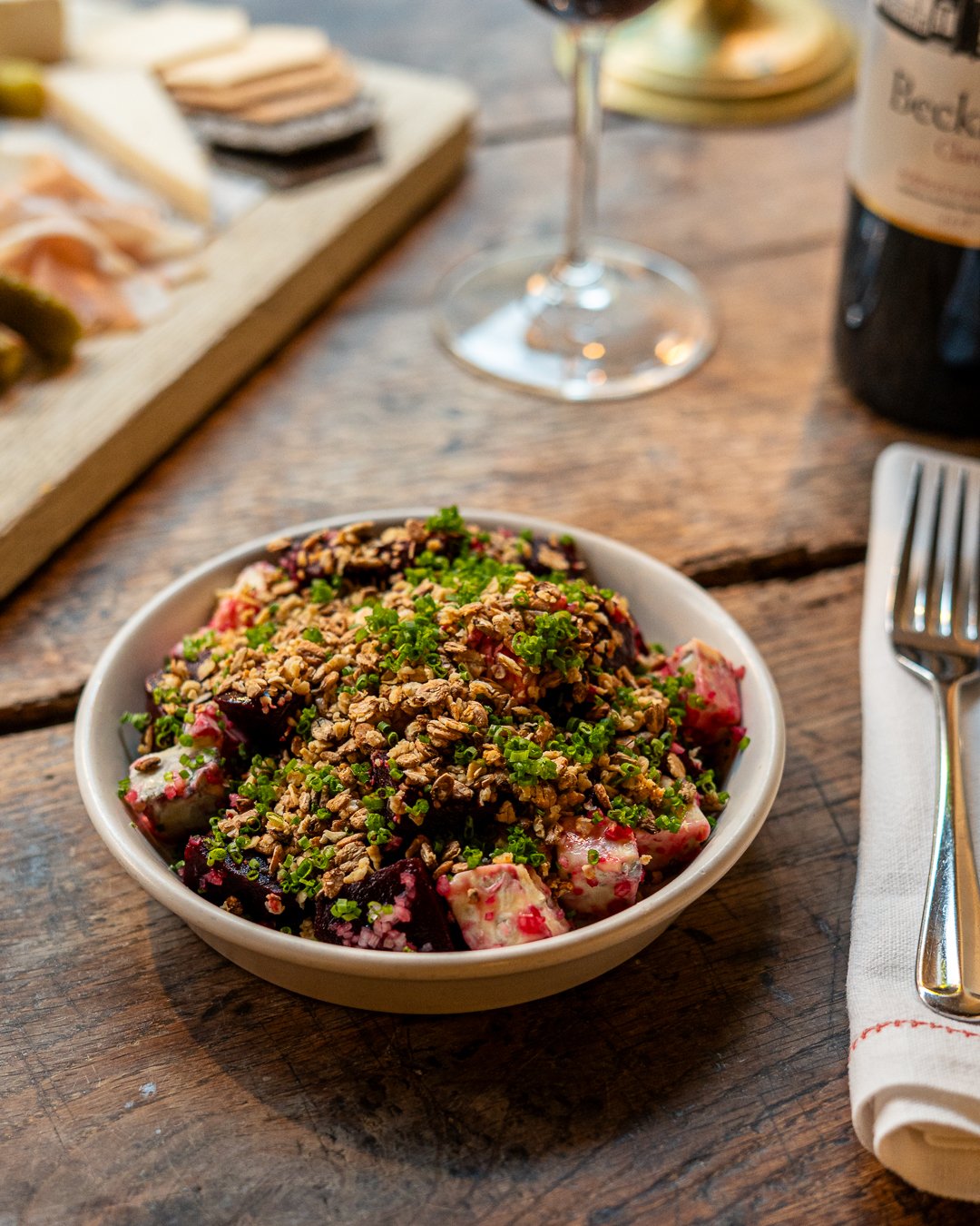 Bowl of berry salad topped with chopped nuts and green herbs on a wooden table with a fork, napkin, wine glass, and bottles in the background.