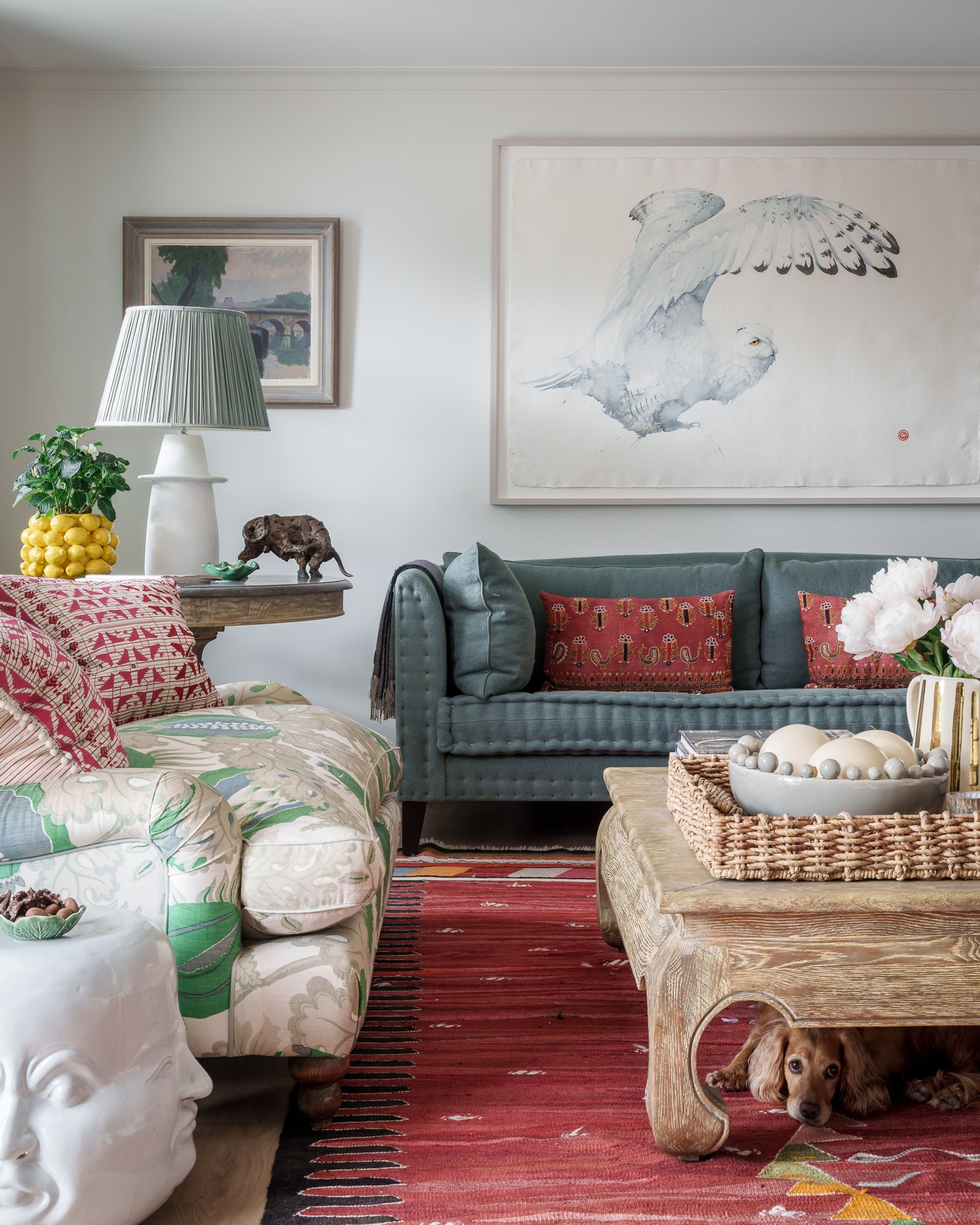 Living room with a large framed feathered bird artwork on the wall, a green velvet sofa with red and black cushions, a wooden coffee table with a decorative bowl of eggs, a lamp on table with a green plant, a painting of a bridge in a frame, and a small dog lying under the table.