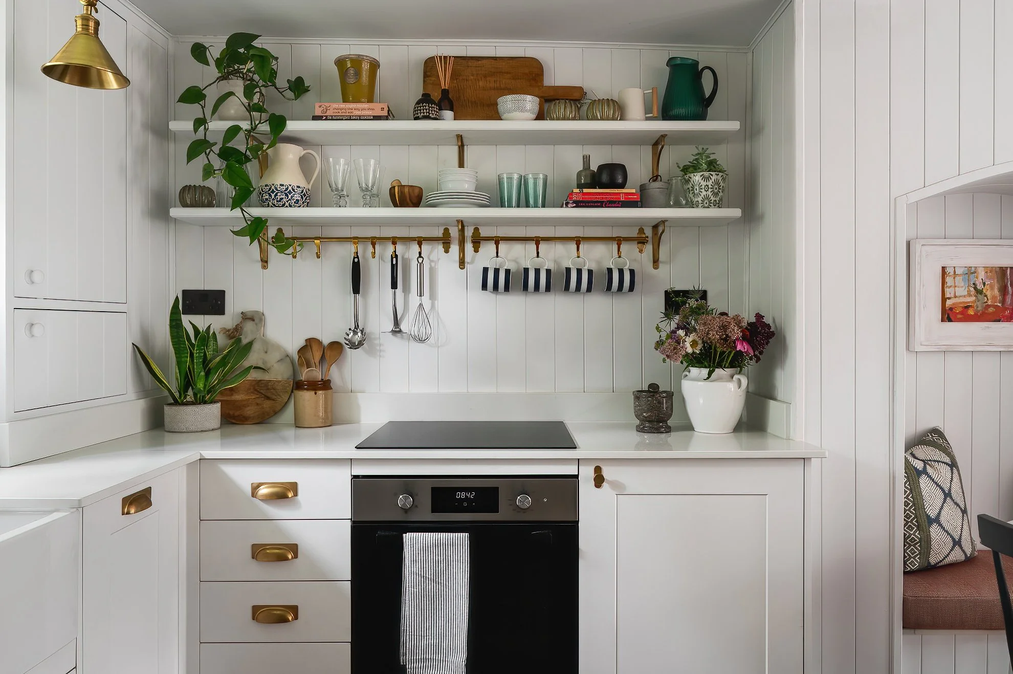 White kitchen with open shelves holding various dishes, bowls, and decorative items. Potted plants and a set of hanging kitchen utensils are also visible.