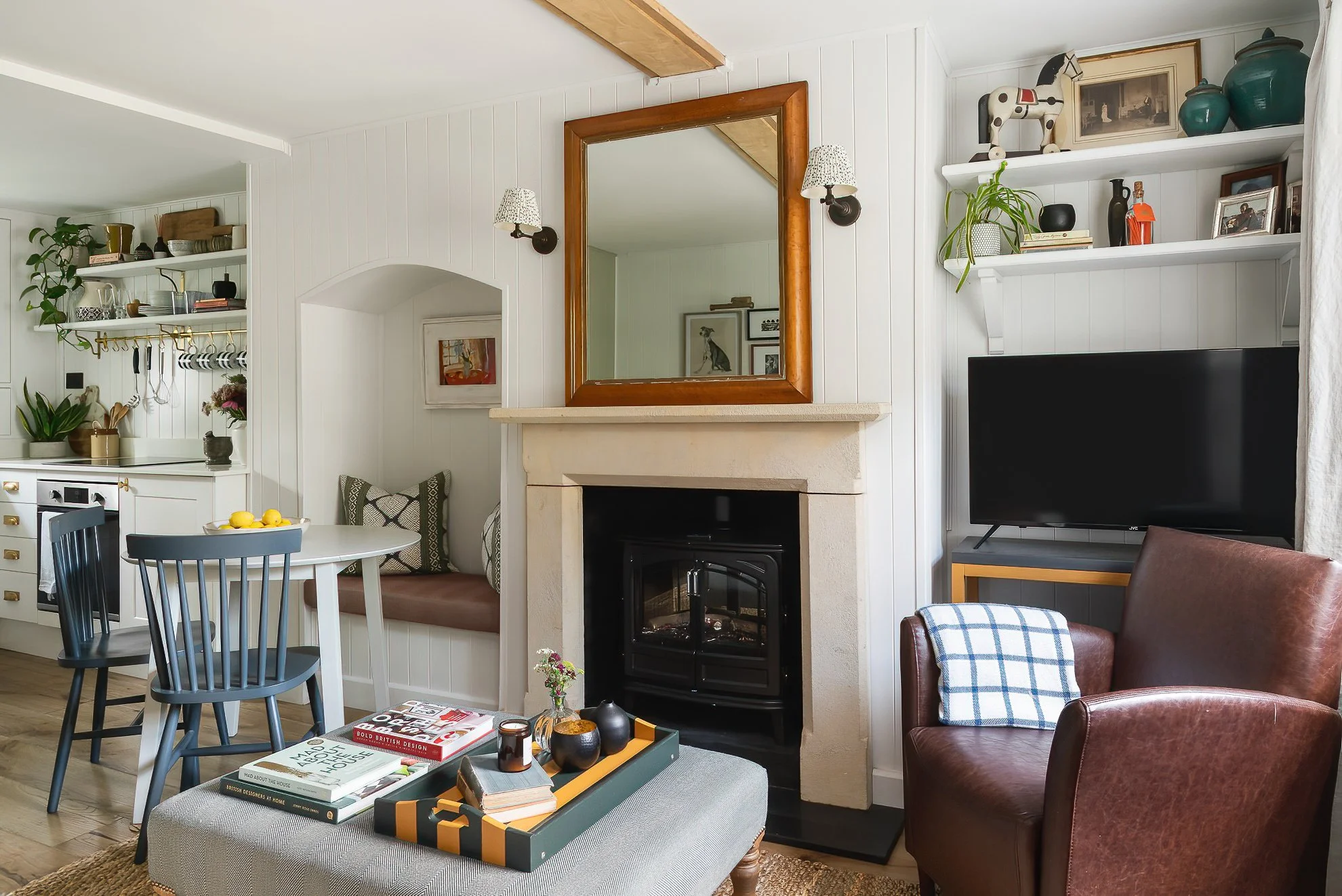 Living room with a fireplace, wall-mounted mirror, shelving with decorative items, and a TV. A round table with chairs and a coffee table with books and decor are visible. A leather armchair with a plaid pillow and a built-in corner bench with cushions are also part of the room.