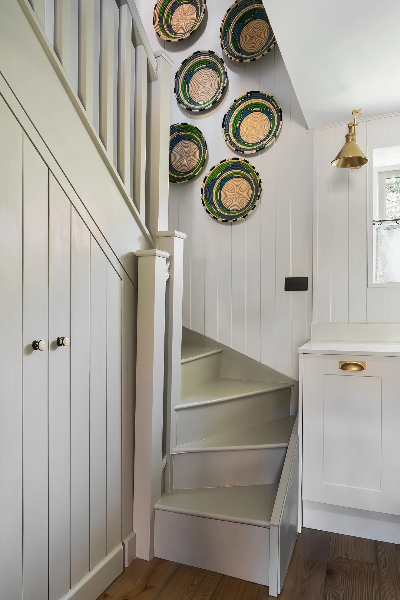 Interior of a house with white wood-paneled walls, a staircase, circular woven baskets on the wall, a brass wall lamp, a small window, and wooden flooring.