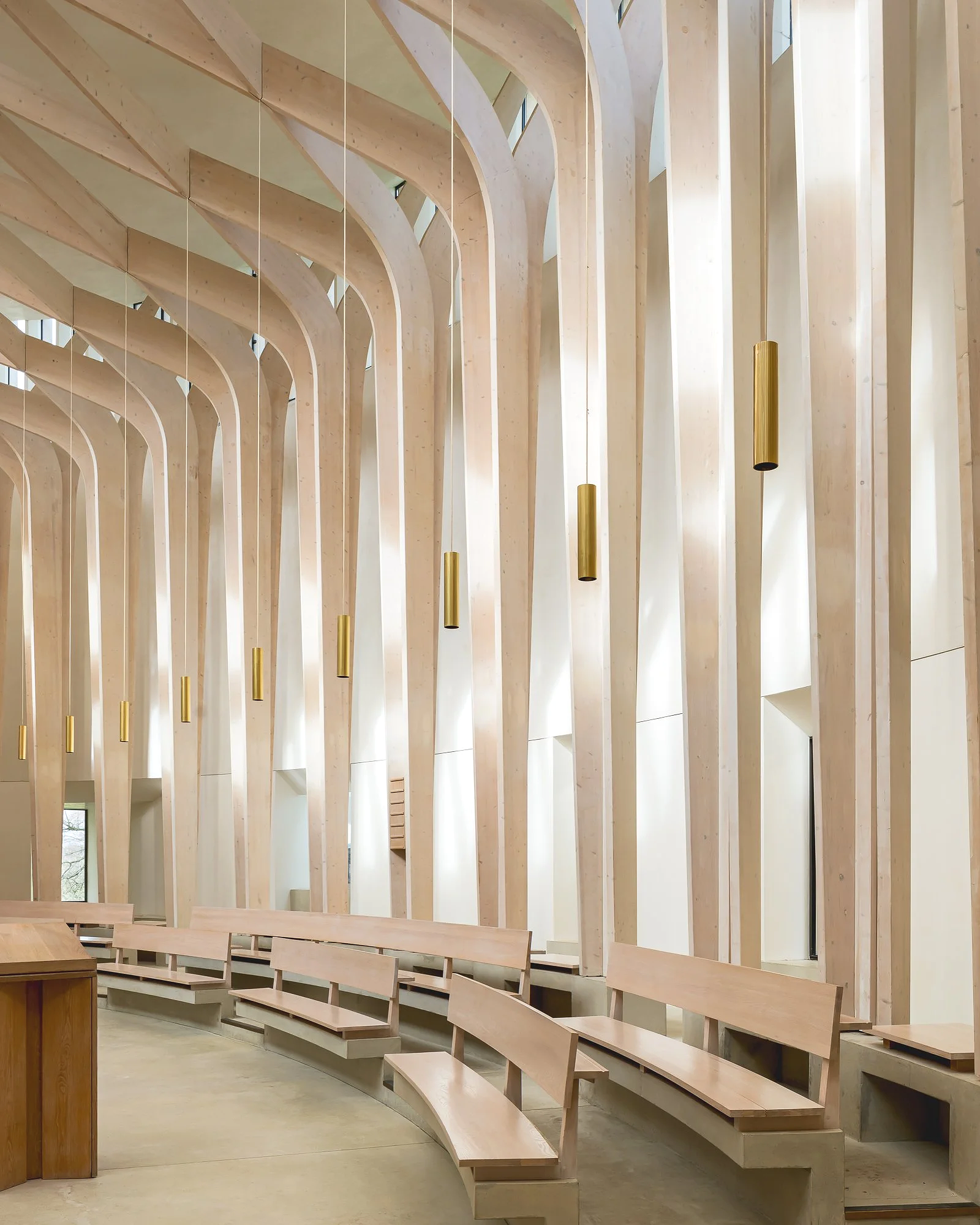 Interior view of a modern building with wooden arches, hanging gold cylindrical lights, and wooden seating arranged in a semicircle.
