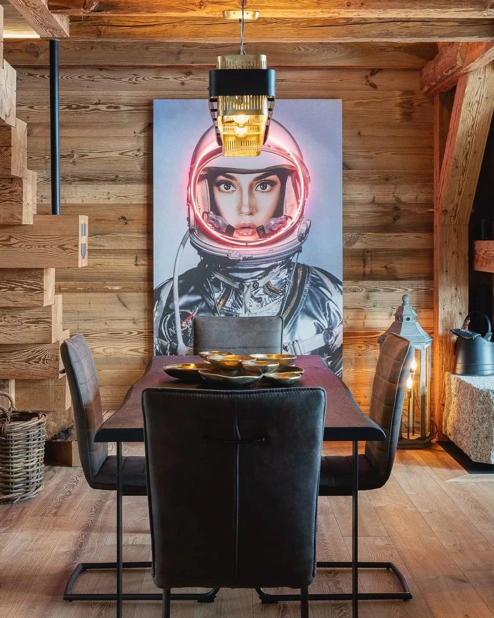 A wooden dining room with a black table and four chairs. A large art piece of a woman in a space helmet with neon lights is on the wall behind the table. There is a pendant light hanging above the table, a wicker basket to the left, and a lantern with candles on the right.