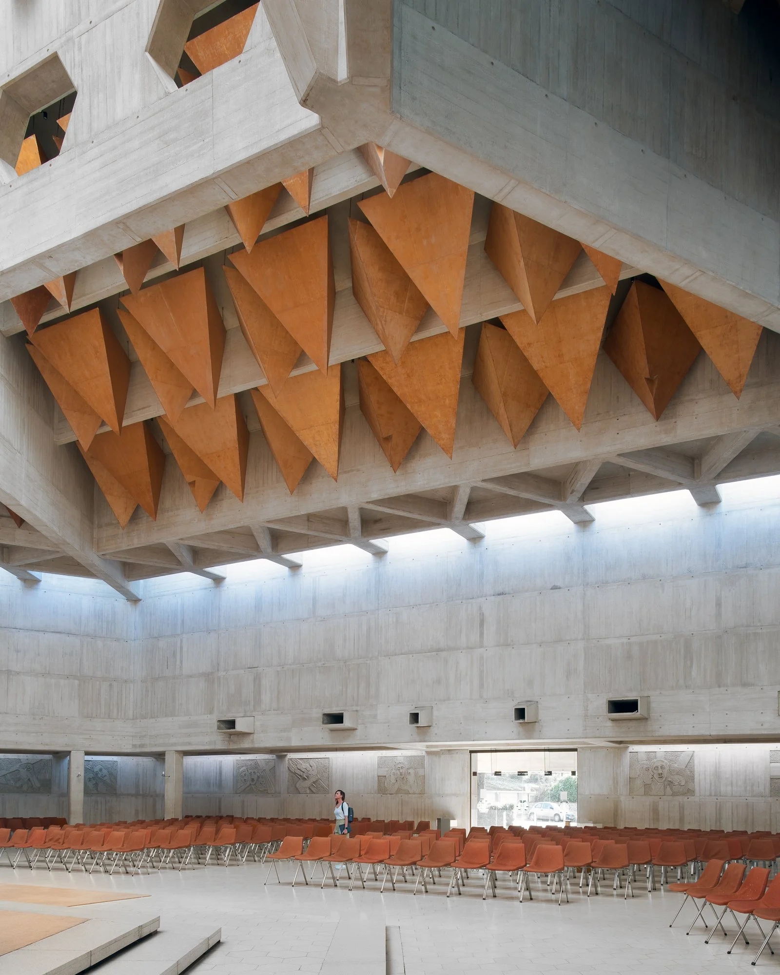 Interior of a modern auditorium or lecture hall with rows of empty orange chairs, a woman standing near the chairs, and a large concrete ceiling with geometric design and orange accents.
