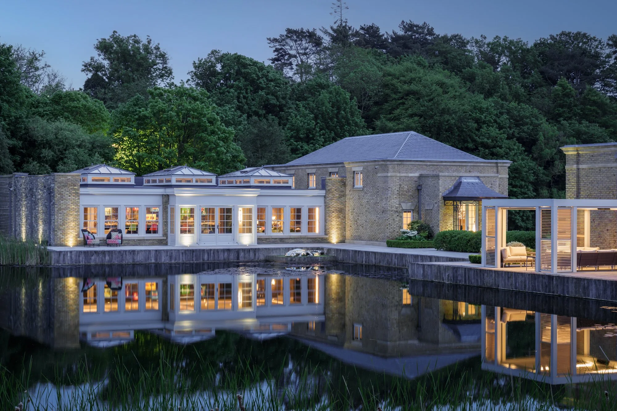 A large house at dusk with bright interior lights, located beside a reflective body of water with a small fountain, surrounded by lush green trees.