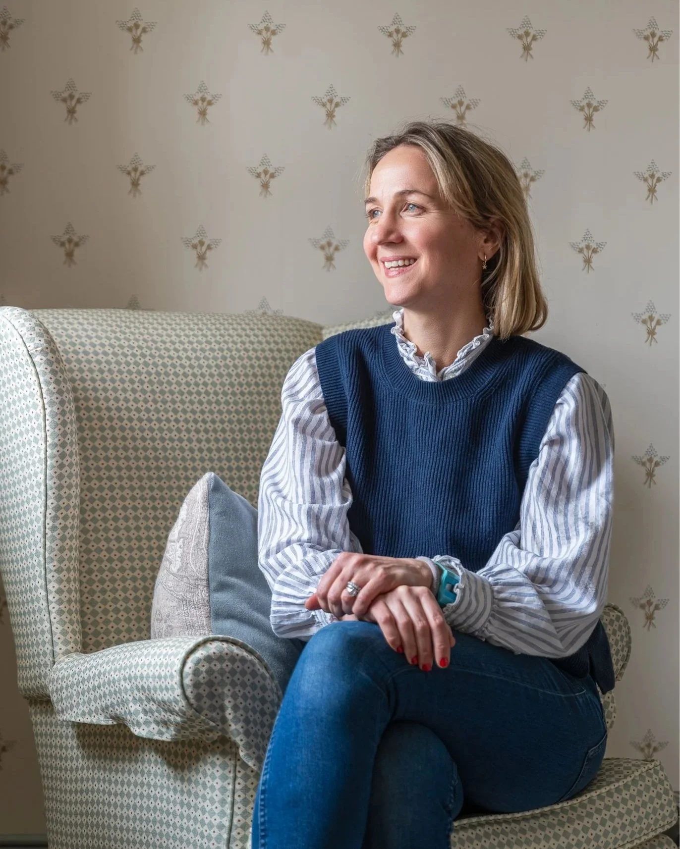 A woman sitting on a patterned armchair smiling, with a floral wallpaper in the background, wearing a blue sweater vest over a striped blouse, jeans, and a watch.