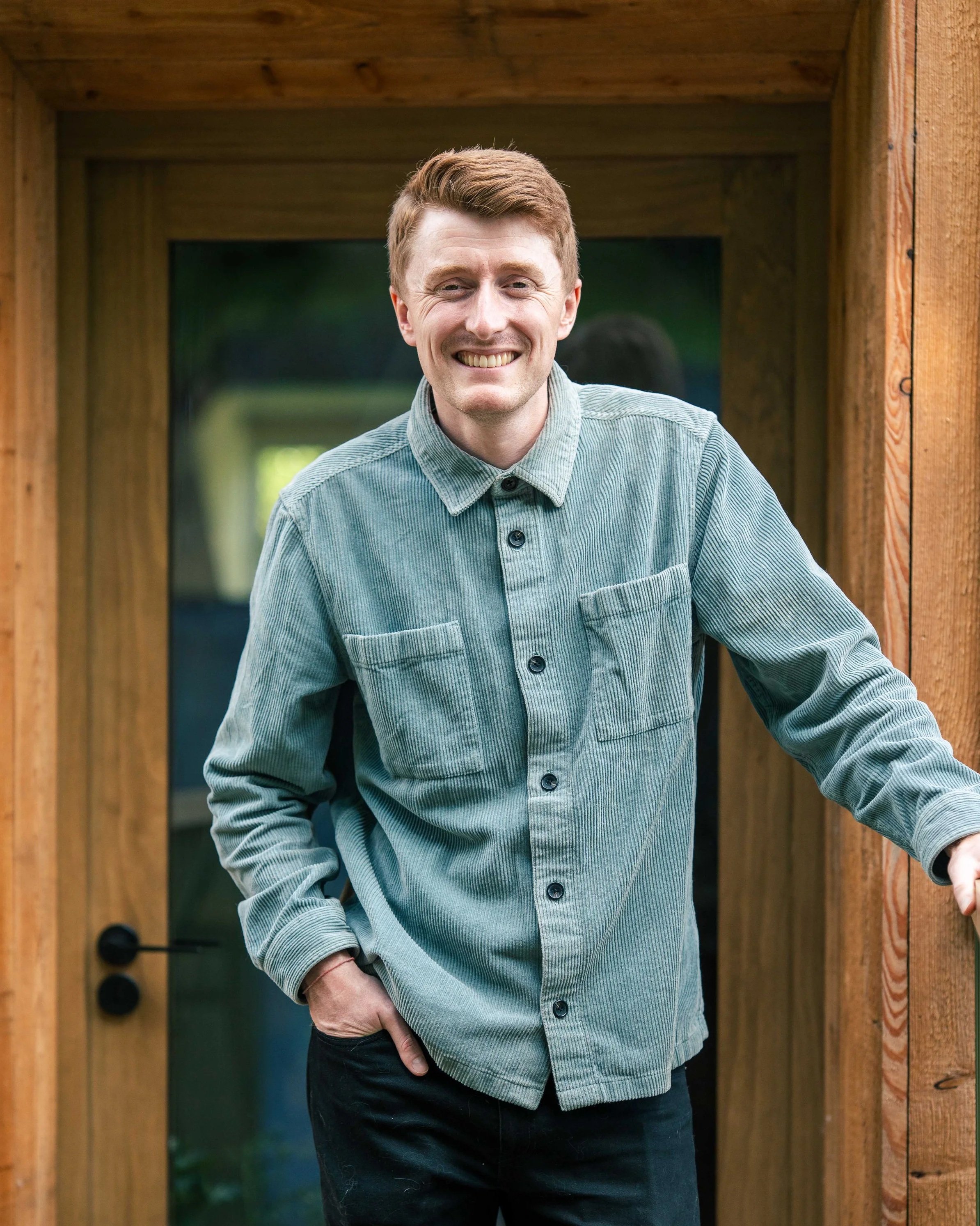 A smiling man with red hair leaning against the doorway of a wooden structure.