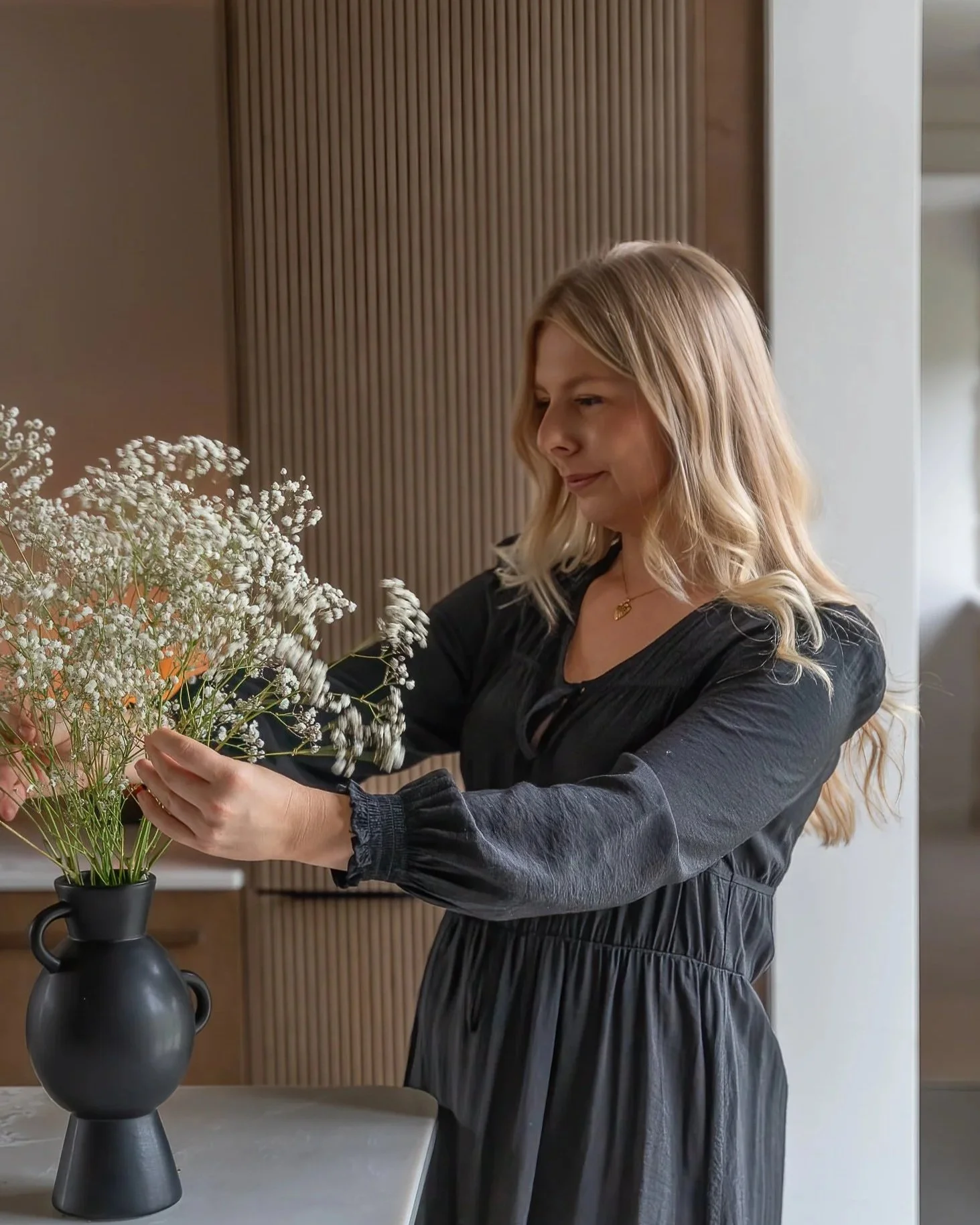 A woman arranging white flowers in a black vase on a kitchen counter.