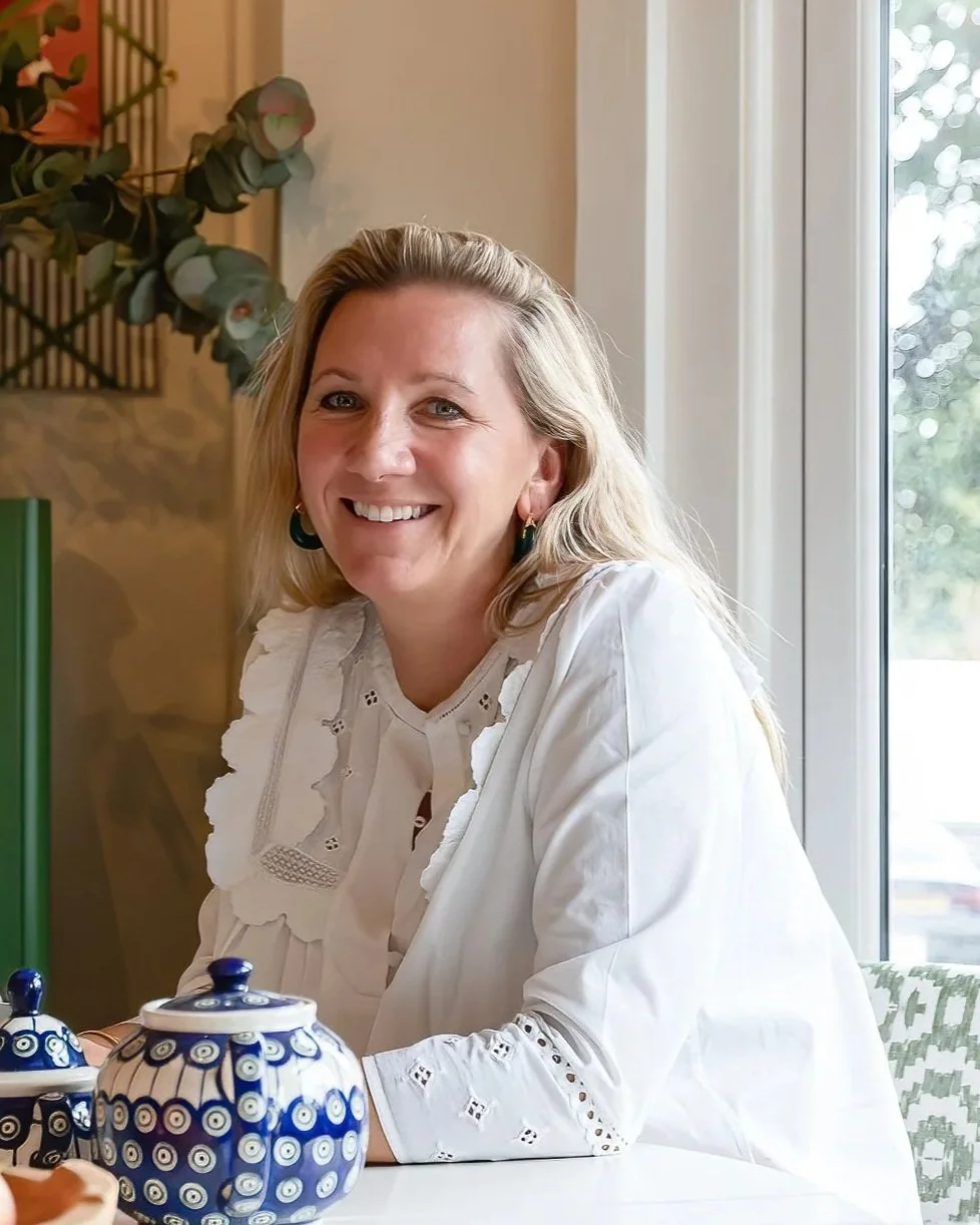 A woman with blonde hair smiling, sitting at a table with blue and white ceramic teapots and cups, near a window in a cozy, well-lit room.