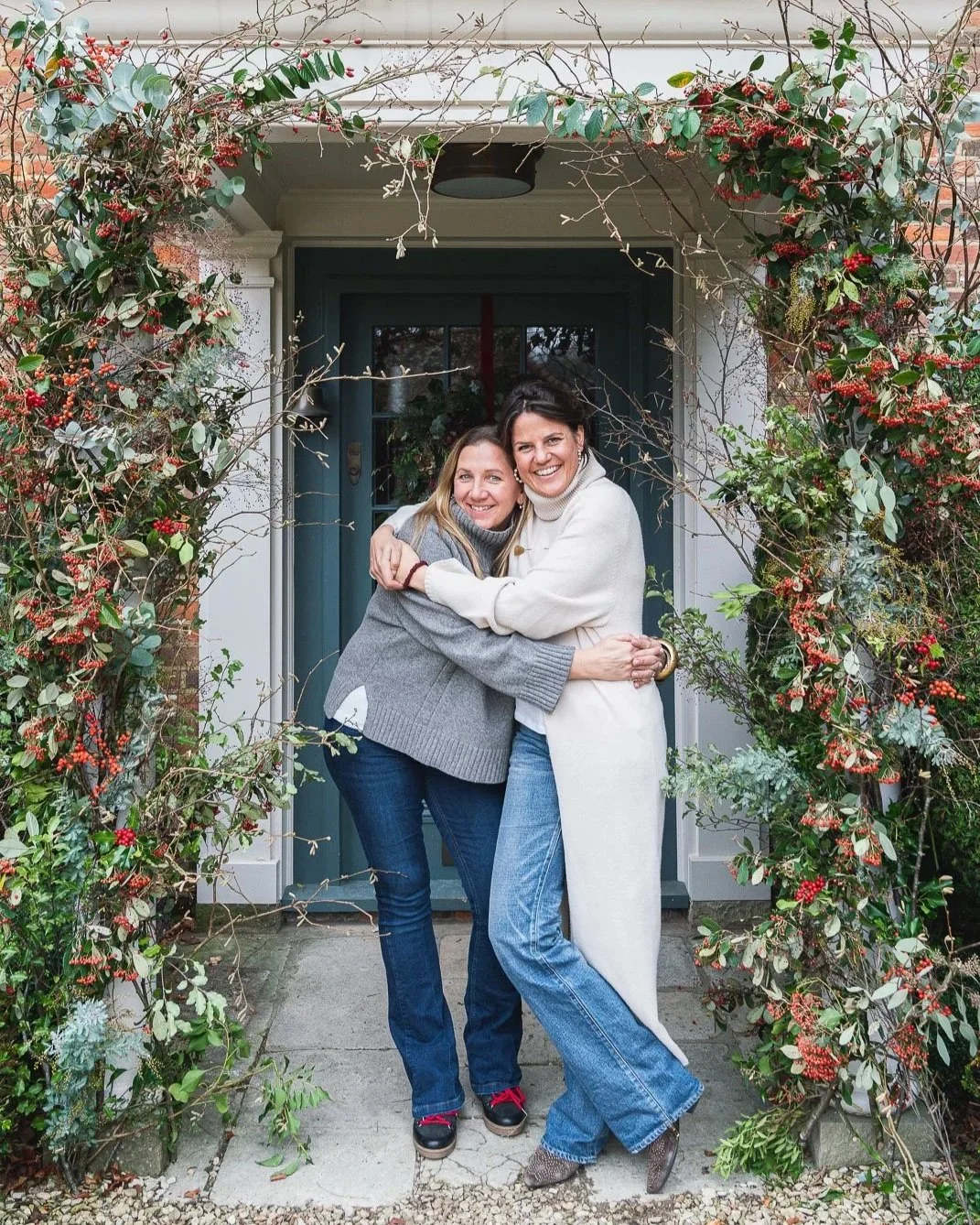 Two women smiling and hugging each other in front of a house entrance decorated with red berries and green foliage.
