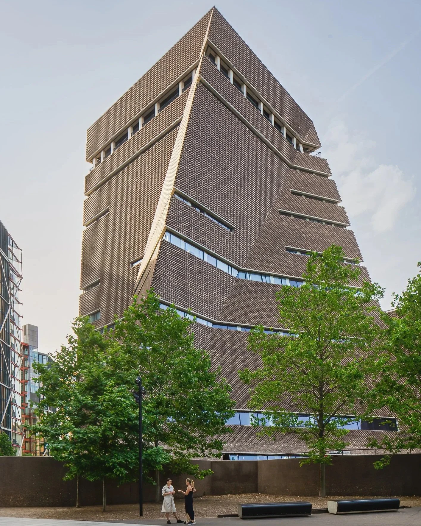 Two women standing and talking in a park with a modern, multi-story building with a brown, textured facade and irregular shape behind them. Trees partially obscure the building.