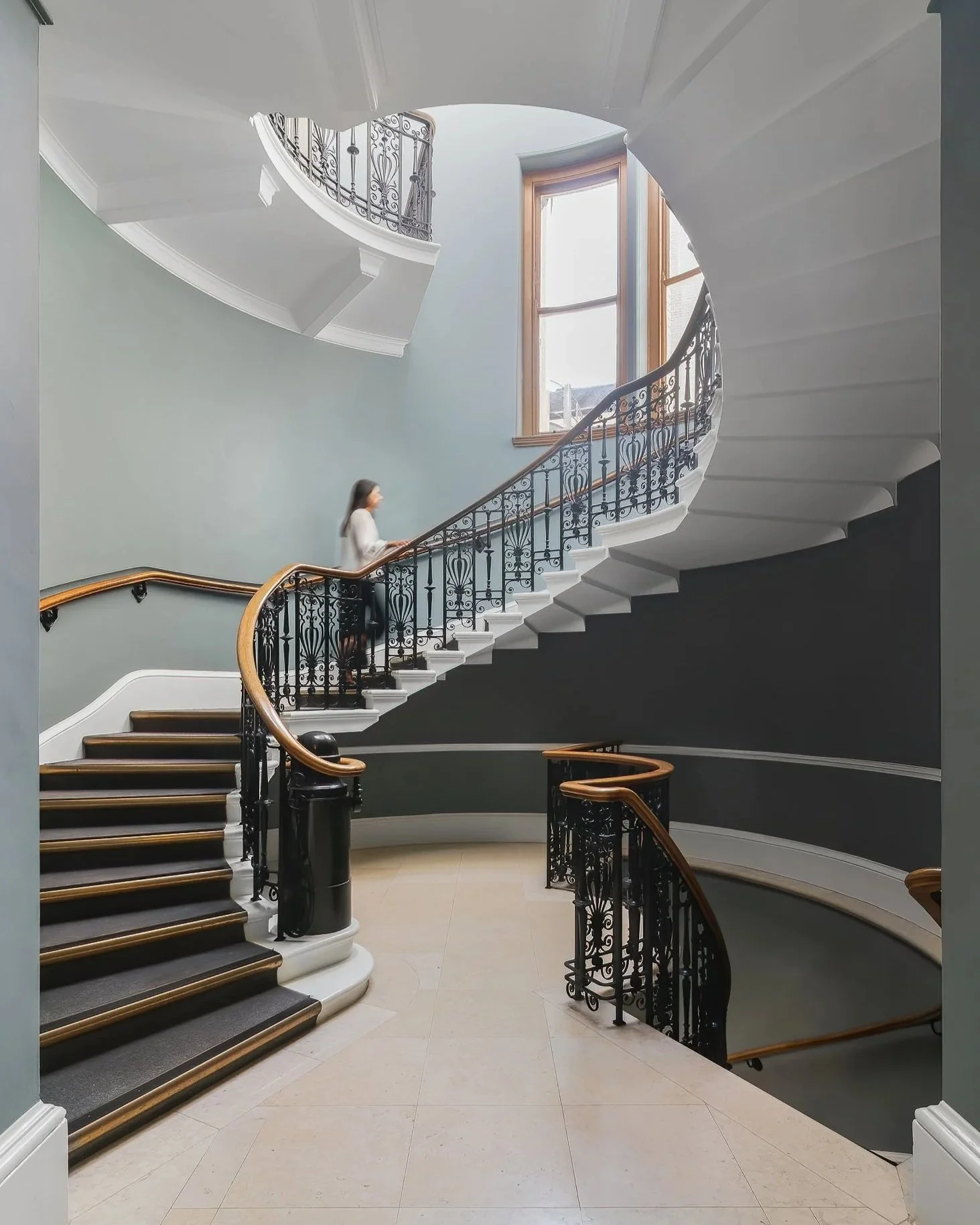 Interior view of a building with a spiral staircase, ornate black railings, wooden handrails, large windows, and a woman walking up the stairs.