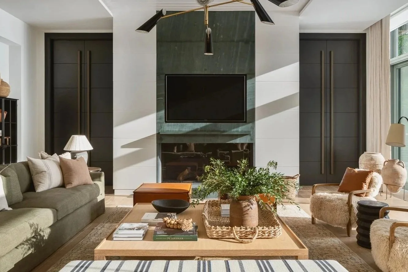 A modern living room with a green sofa, armchairs with sheepskin coverings, a wooden coffee table with books and decorative items, a black cabinet with vases, a ceiling fan with light fixtures, a fireplace, and a large window with curtains overlookin