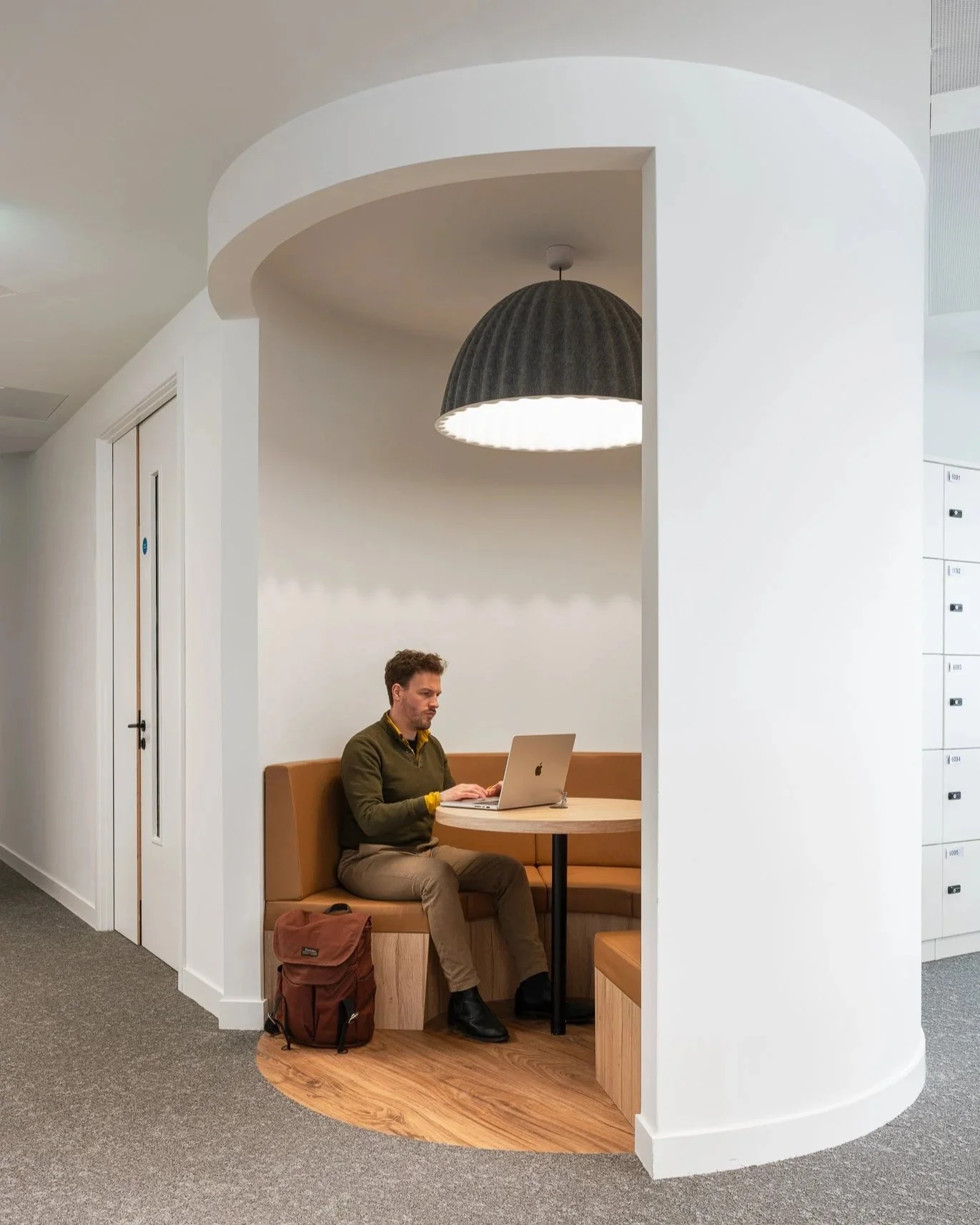 A man sitting in a semi-circular booth working on a laptop in a modern, minimalist office space.
