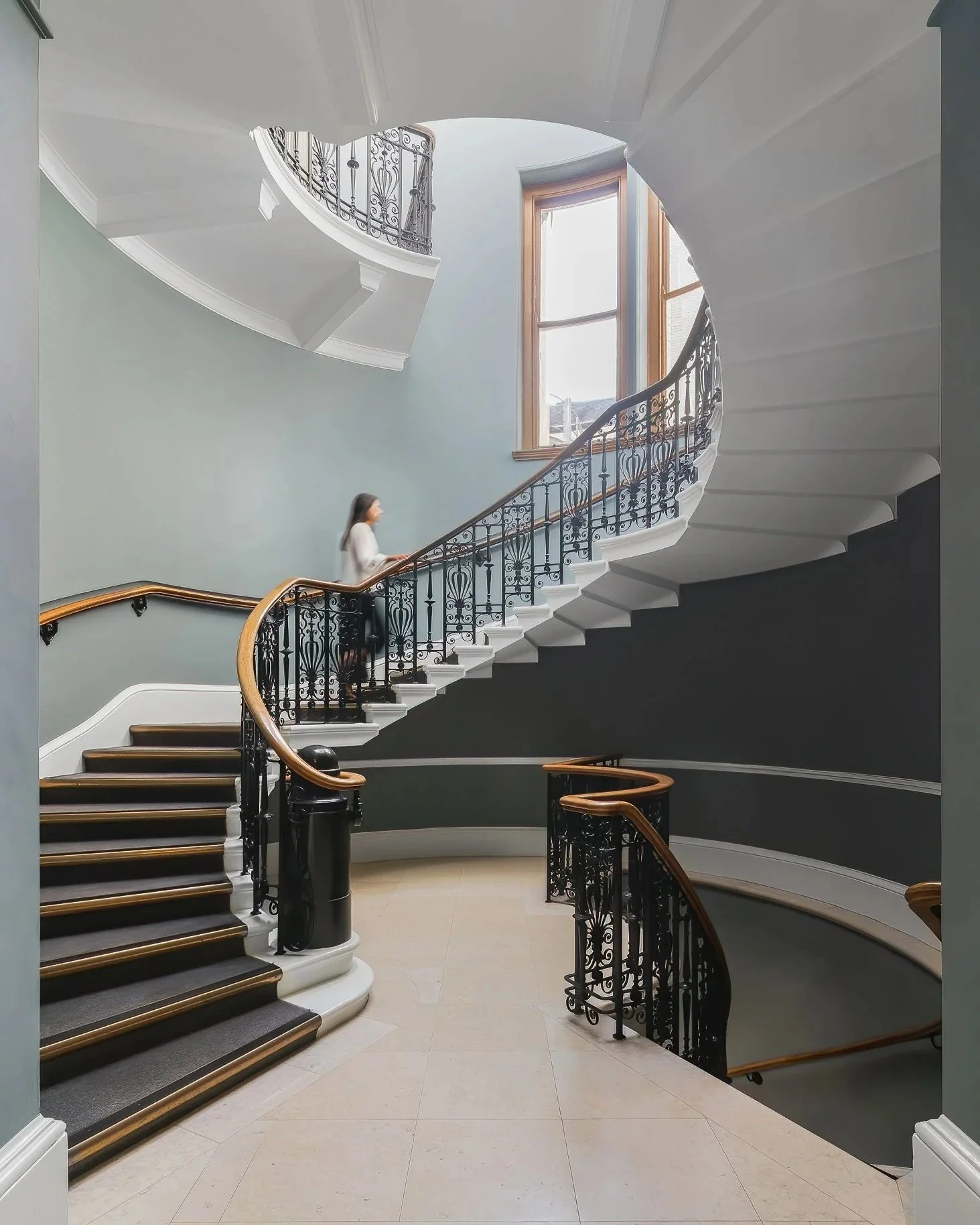 An elegant spiral staircase with black ornate metal railing, wooden handrails, leading to upper floors, with a woman walking up the stairs inside a building with large windows and light-colored walls.