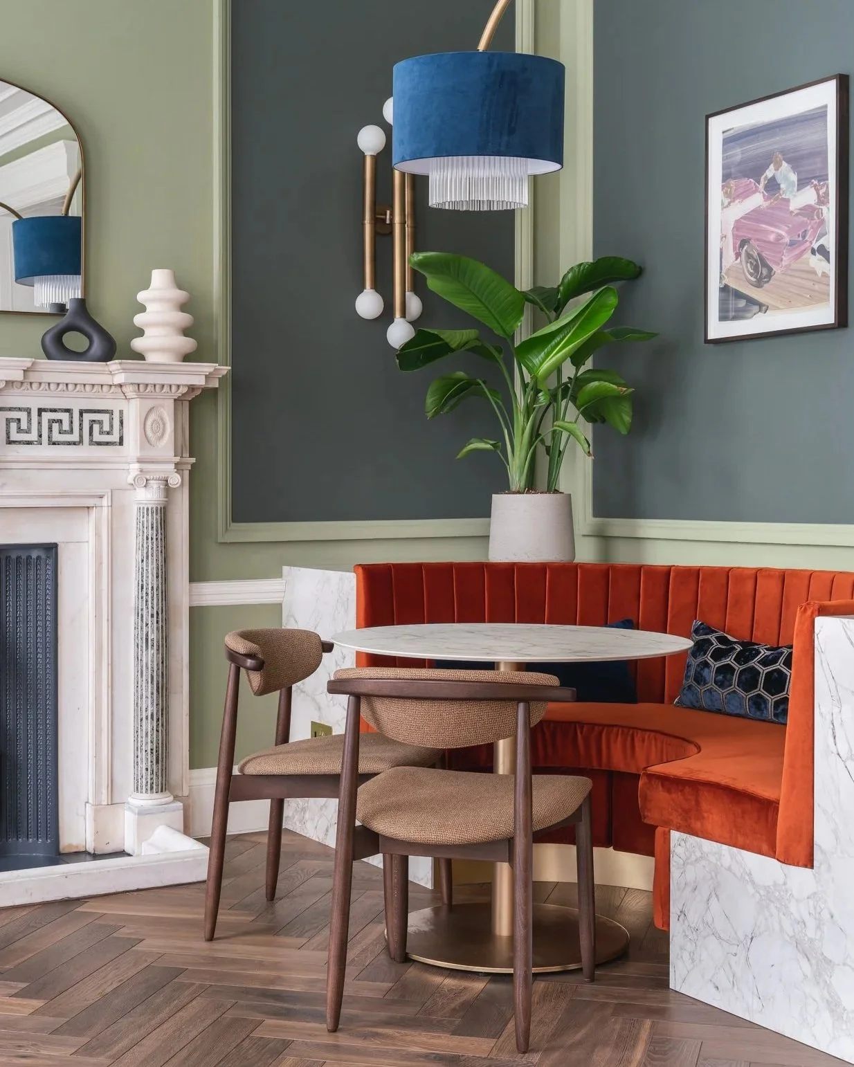 Interior living space with green and dark blue wall panels, a white marble fireplace with decorative objects on mantel, a potted plant, an orange velvet banquette, a round marble table, and two wooden chairs with beige upholstery.