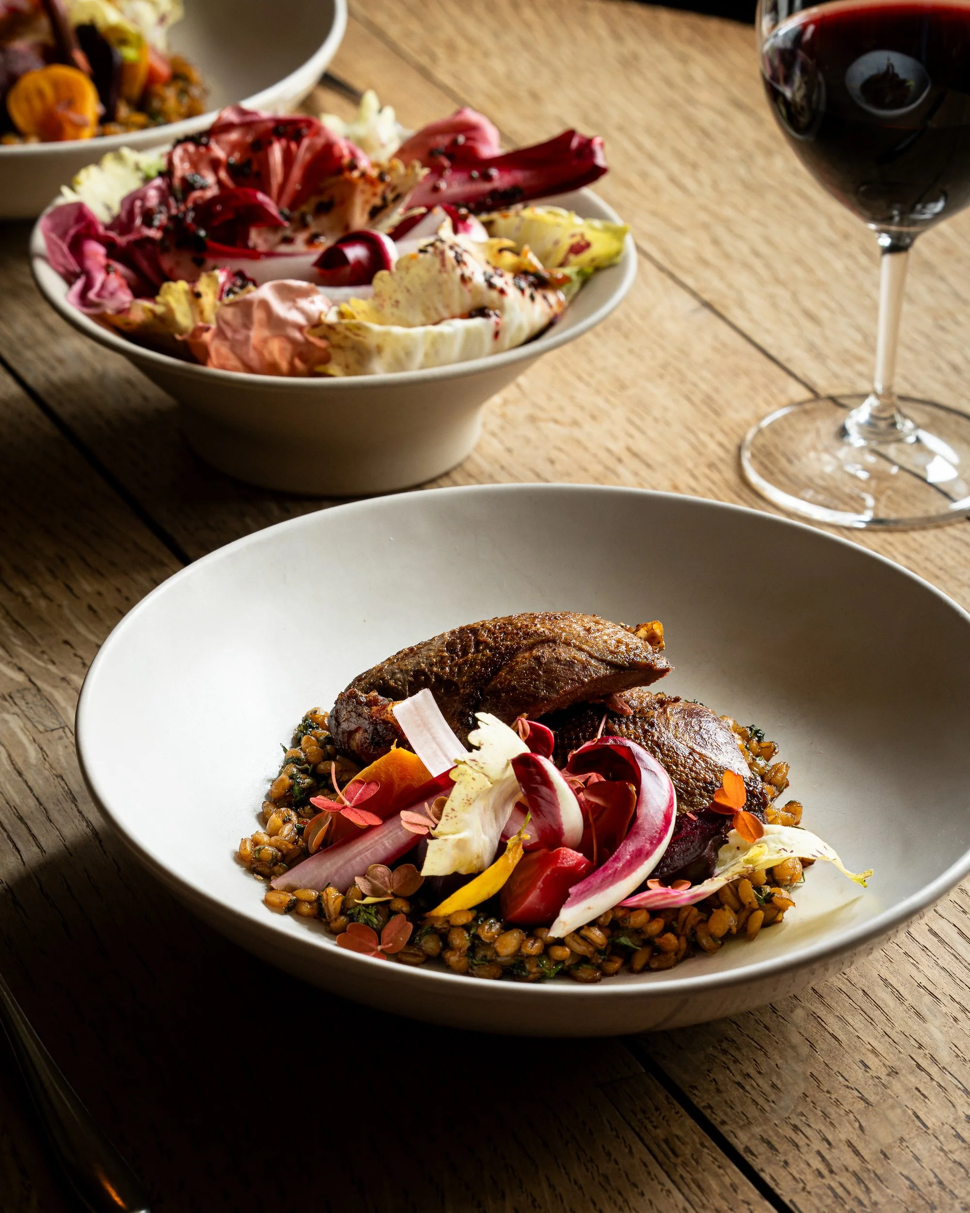 A plated dish features a piece of cooked meat, likely duck, served on a bed of lentils with colorful edible flower petals and pickled vegetables. A glass of red wine is in the background on a rustic wooden table.