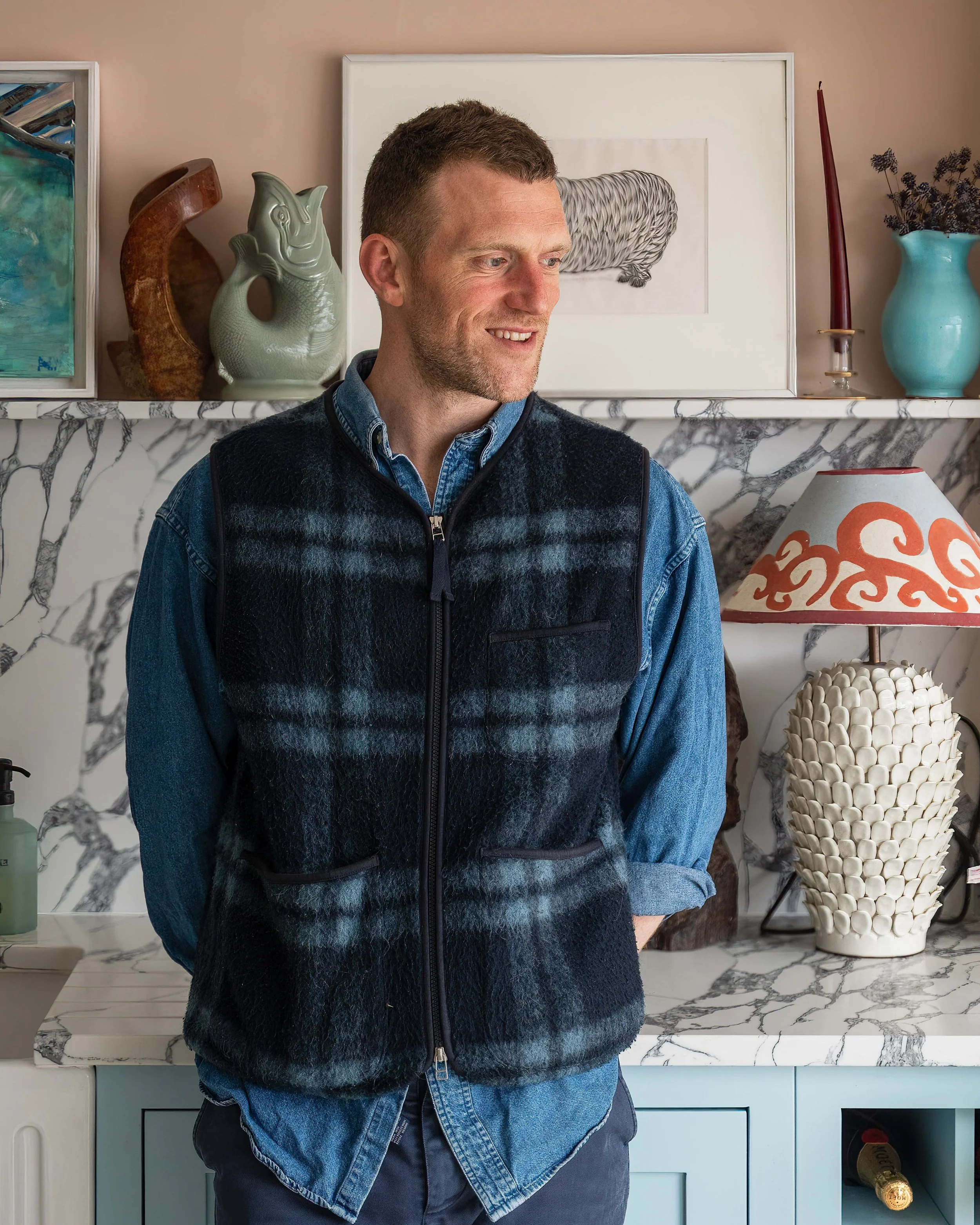 A man standing in a room with decorative items on a marble countertop and shelves, including vases, artwork, and a lamp, wearing a blue denim shirt and a black fleece vest.