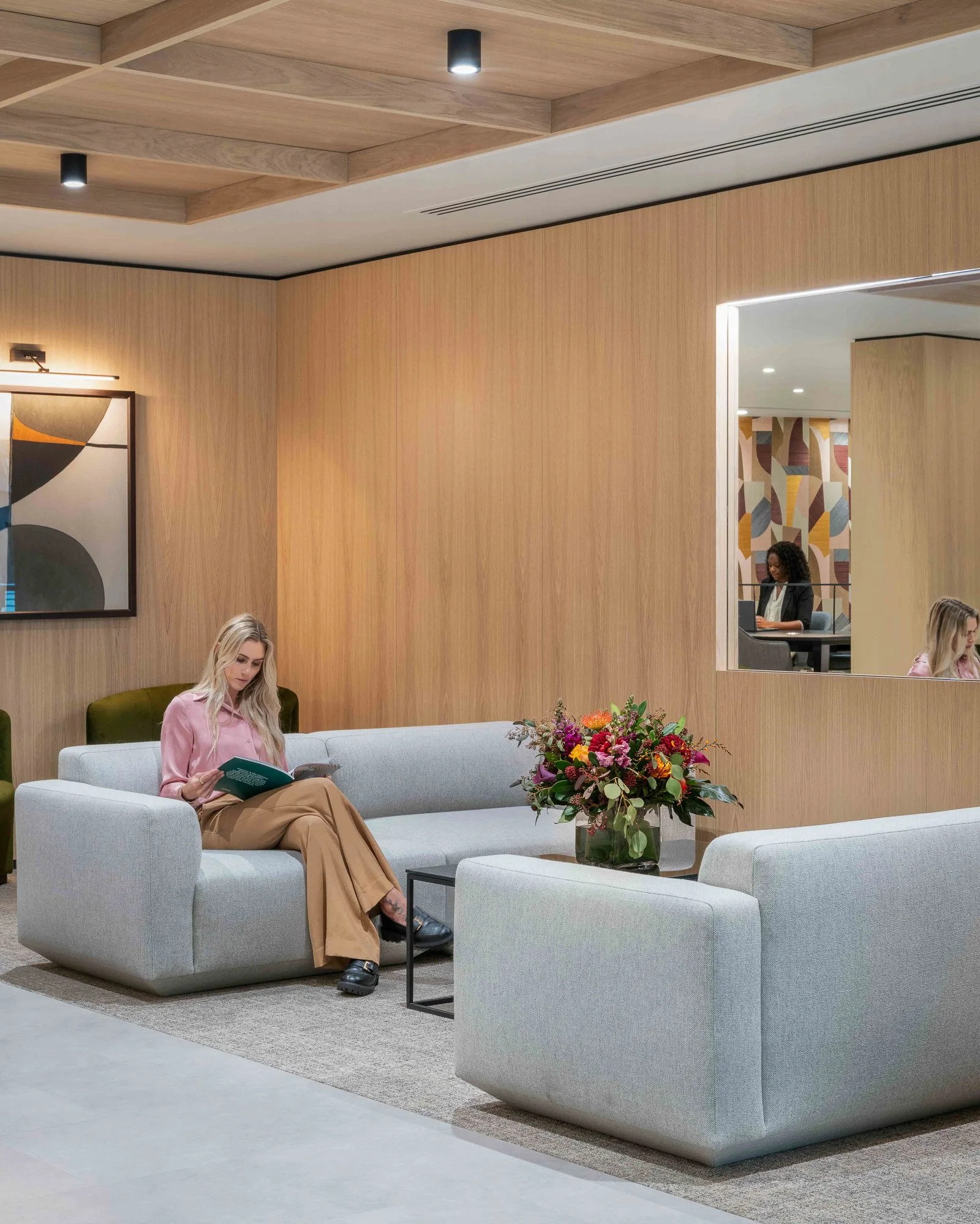 A modern lounge area with light gray couches, a glass side table, a large flower arrangement, wooden wall paneling, and a mirror reflecting two women sitting at a table in the background.