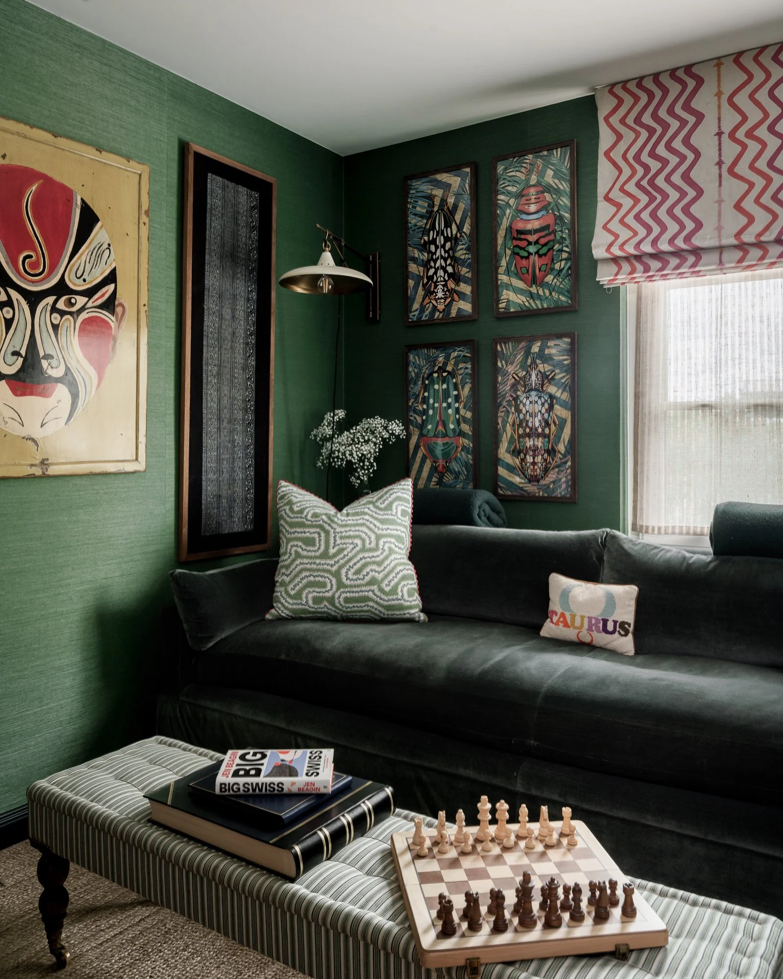 Living room corner with a dark green sofa, patterned pillows, an abstract art piece, and a gallery wall of insect illustrations. A white and black striped ottoman with books and a chessboard is in front, near a window with red and white striped blinds.