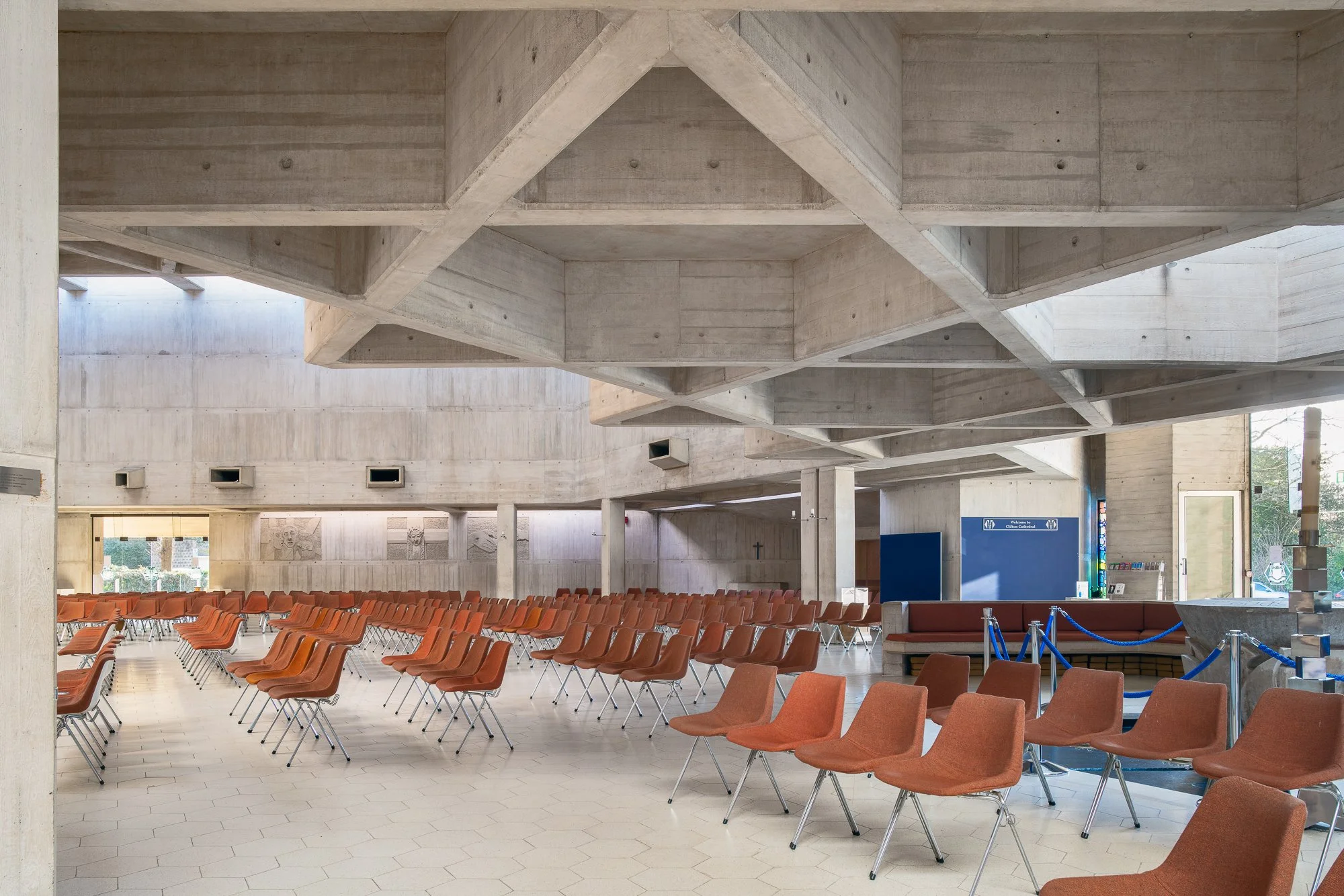 Interior of a large modern building with concrete architecture, rows of orange chairs, and a high ceiling with geometric concrete designs.