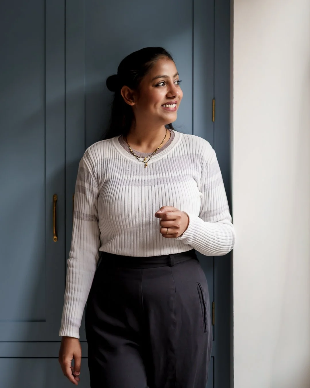 A young woman with dark hair tied back, smiling and looking to her right, standing indoors against blue cabinet doors, wearing a white ribbed sweater, dark high-waisted pants, gold necklace, and a ring on her left hand.
