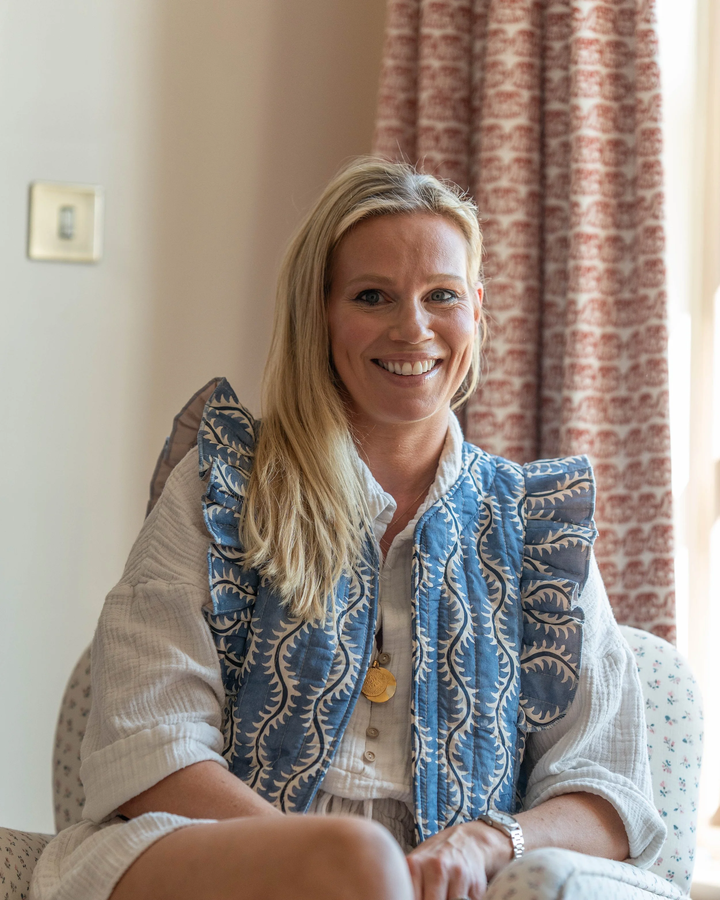 A smiling blonde woman sitting indoors in front of a window with pink patterned curtains, wearing a beige top with blue embroidered ruffles and accessorized with layered necklaces and a watch.