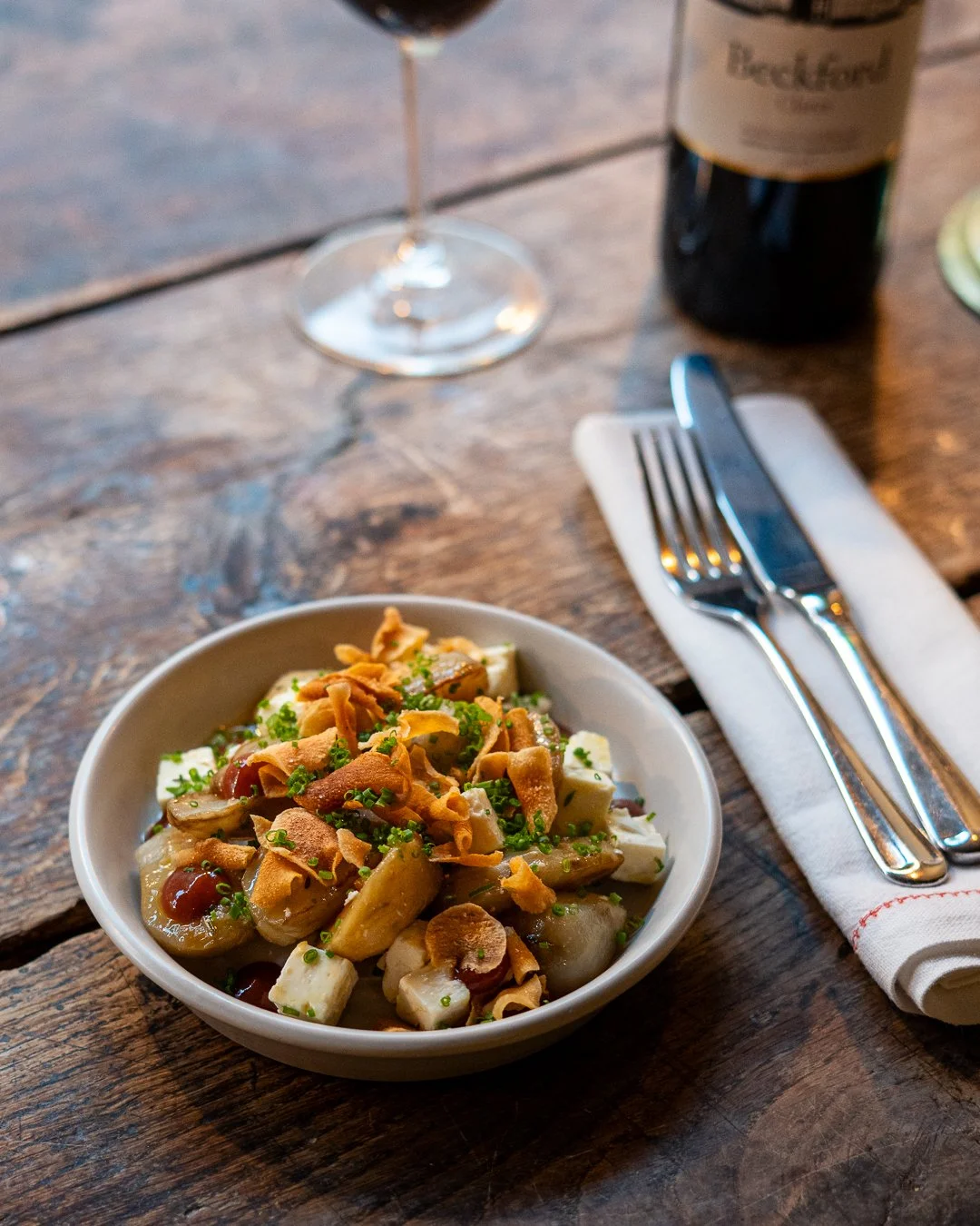 A bowl of artichoke salad topped with chopped chives and crispy fried onions on a rustic wooden table, with a glass of red wine, a bottle of wine, and silverware on a napkin nearby.