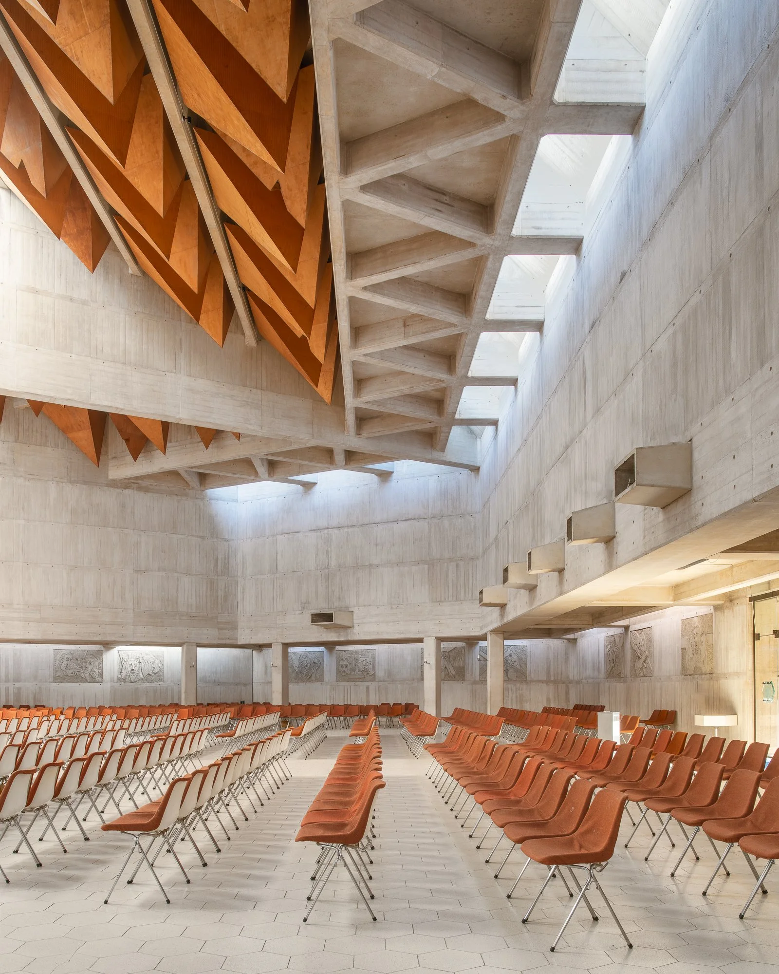 An empty auditorium with rows of orange chairs, a large concrete wall with decorative bas-reliefs, and a high ceiling with natural light coming from skylights.