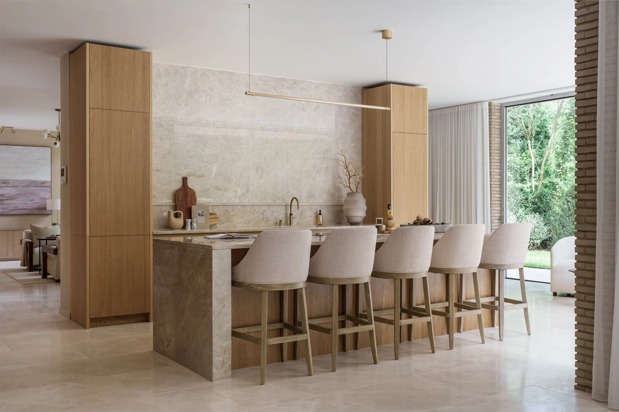 A modern kitchen and dining area with a large wooden counter and high stools, beige and neutral tones, decorative vases, and large windows showing green trees outside.
