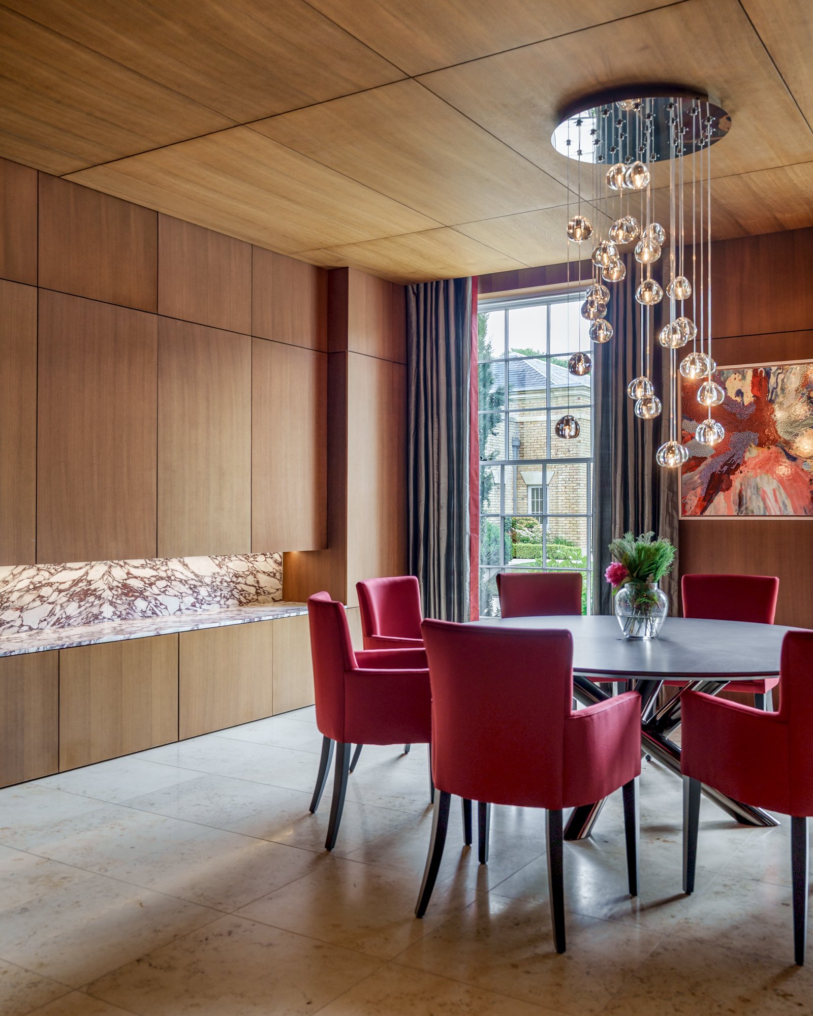 A modern dining room with a round dining table surrounded by red upholstered chairs, large windows with dark curtains, a colorful abstract painting, a contemporary chandelier, and a marble sideboard with a wooden wall behind it.