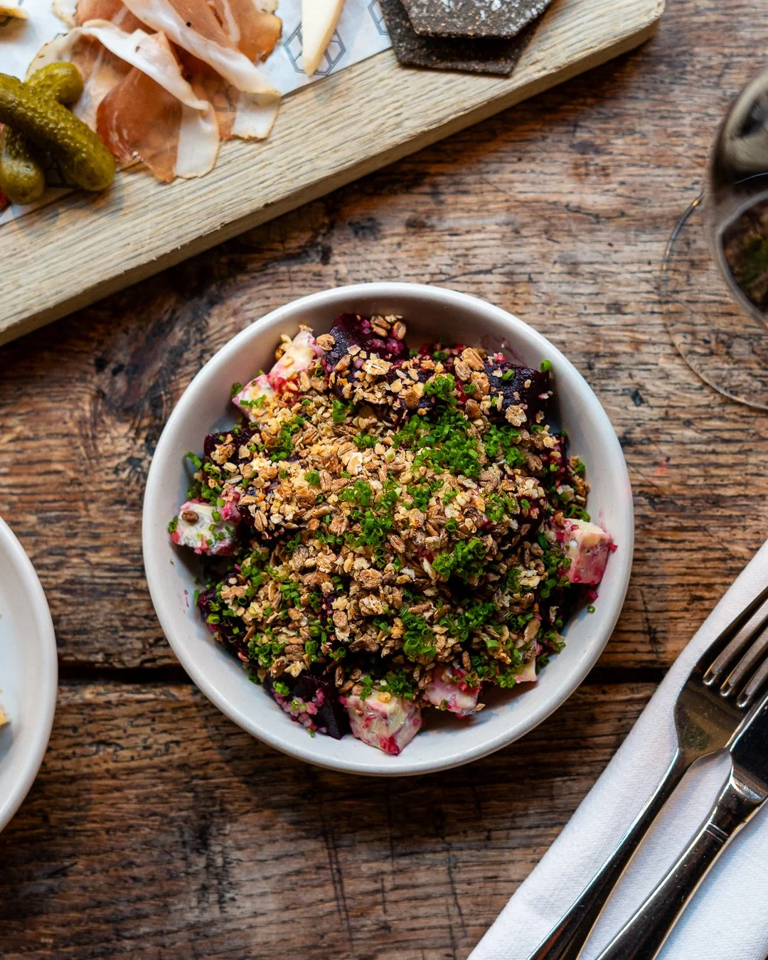 A bowl of beet salad topped with chopped nuts and chopped green herbs, on a rustic wooden table.