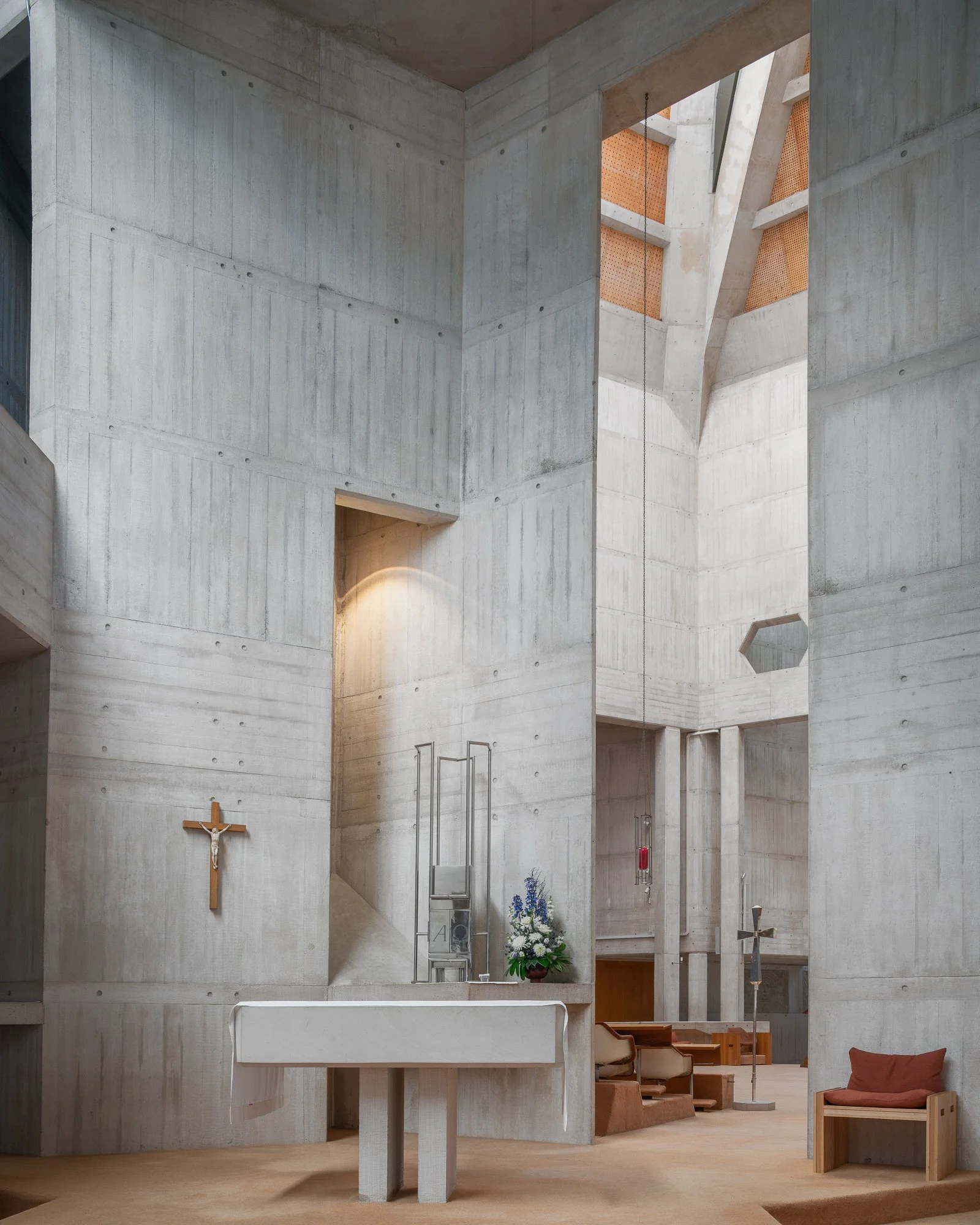 Interior of a modern church with concrete walls, a wooden crucifix on the left, a flower arrangement, seating, and religious symbols.