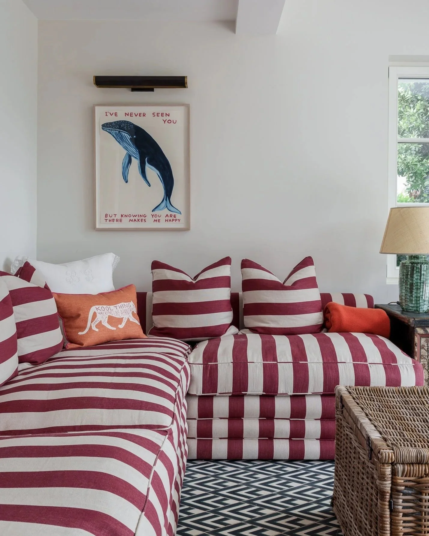 Living room with striped red and white sofa and pillows, a wicker basket, a lamp, a window with greenery outside, and wall art of a whale with text.