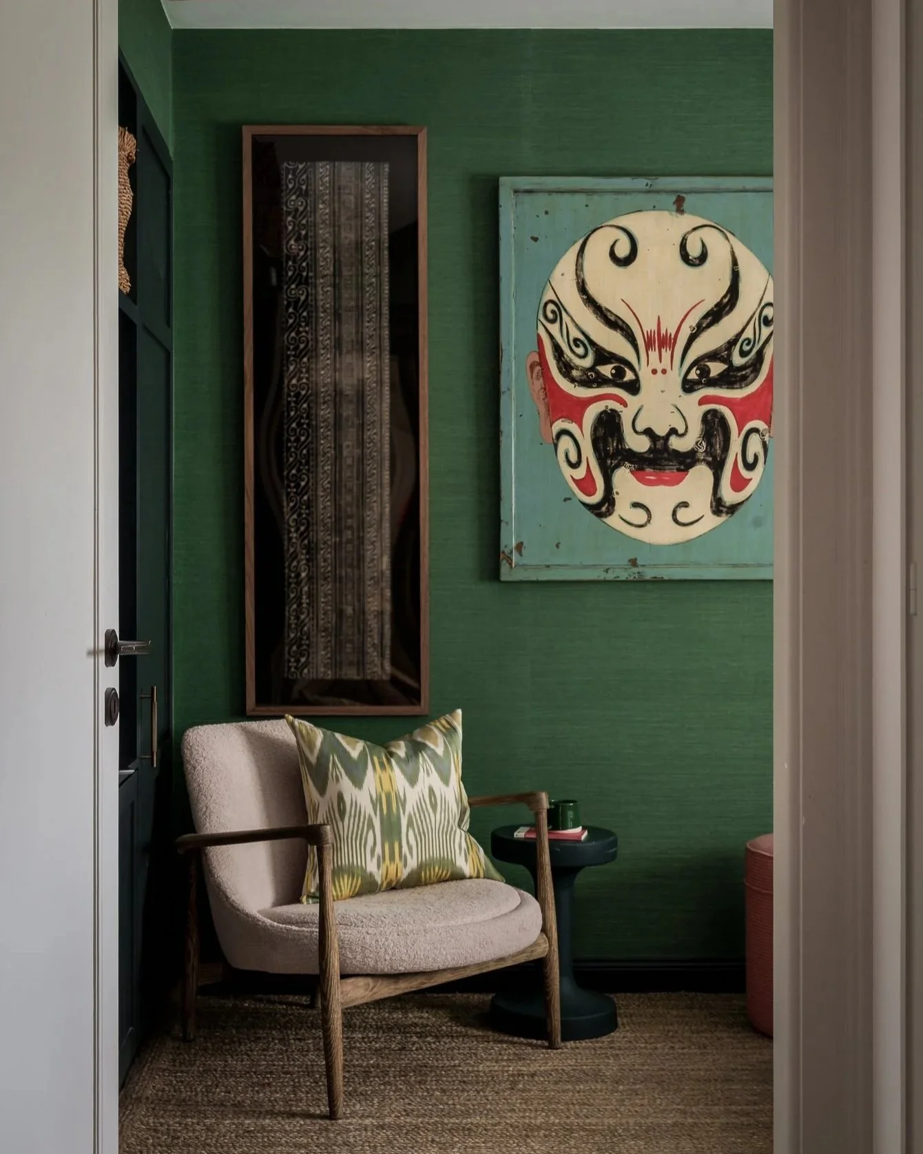 A cozy corner with a beige upholstered armchair featuring wooden armrests and a decorative cushion with green and white geometric patterns. A small round black side table holds a green cup and various items. The background includes a green textured wall, a wooden-framed mirror, and a colorful face mask artwork with bold black, red, and white patterns.