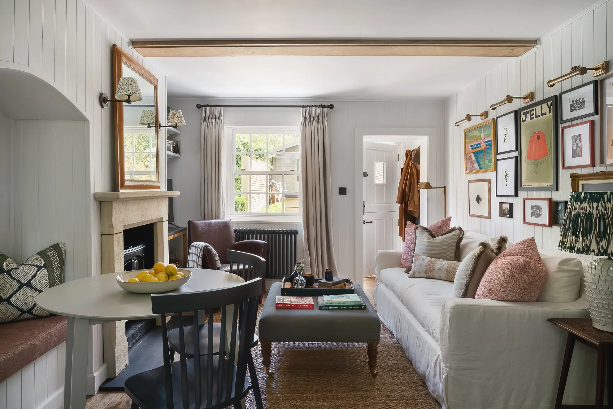 A cozy living room with white paneled walls, a white sofa with patterned and solid cushions, a dark wooden coffee table with books, framed pictures, and decorative objects, a dining table with lemon fruit bowl, beige curtains, and a picture gallery wall near the ceiling. Windows show greenery outside.