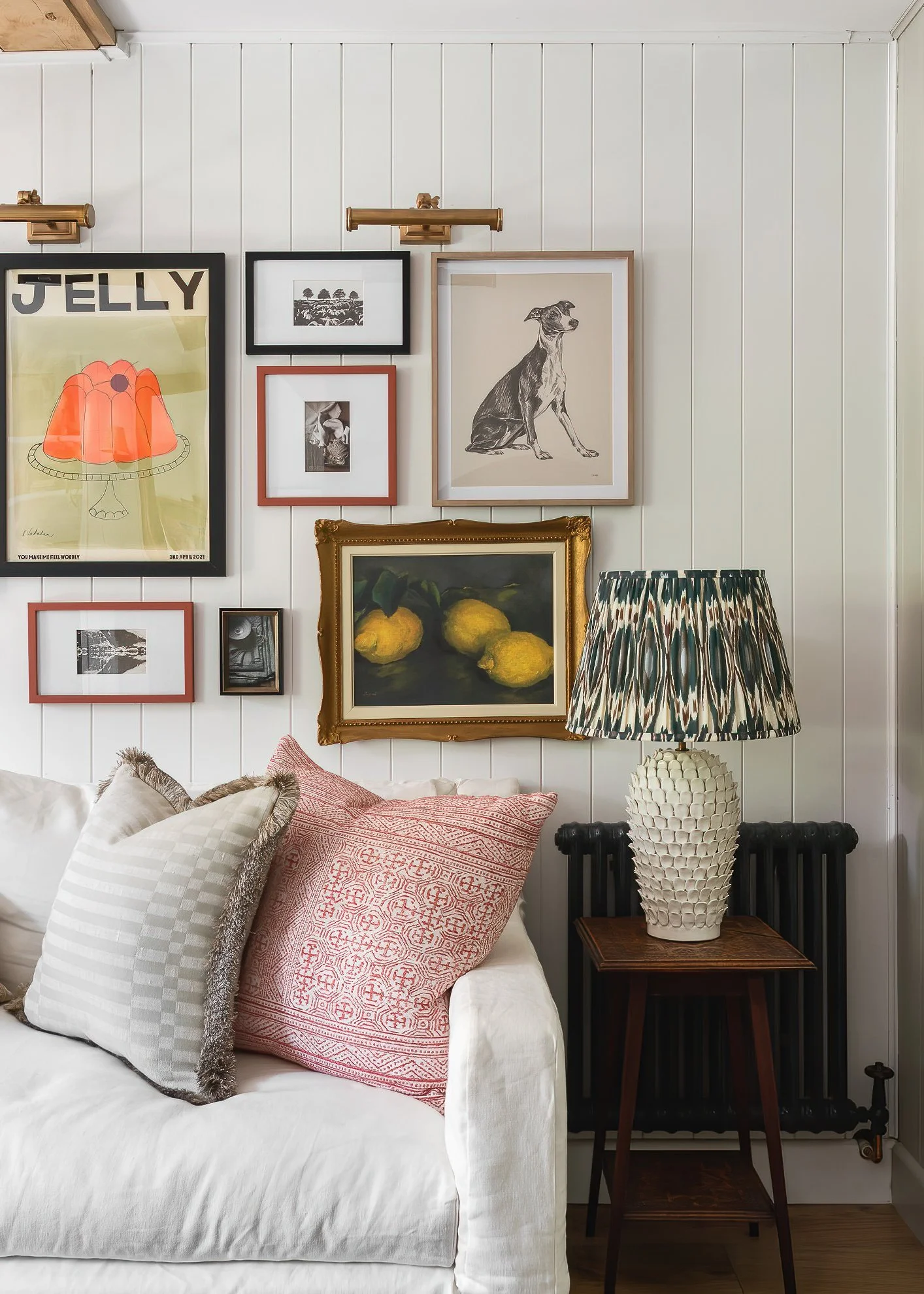 A white bed with multiple patterned pillows, a wall with framed art and photos, a vintage lamp, and a decorative vase on a side table.