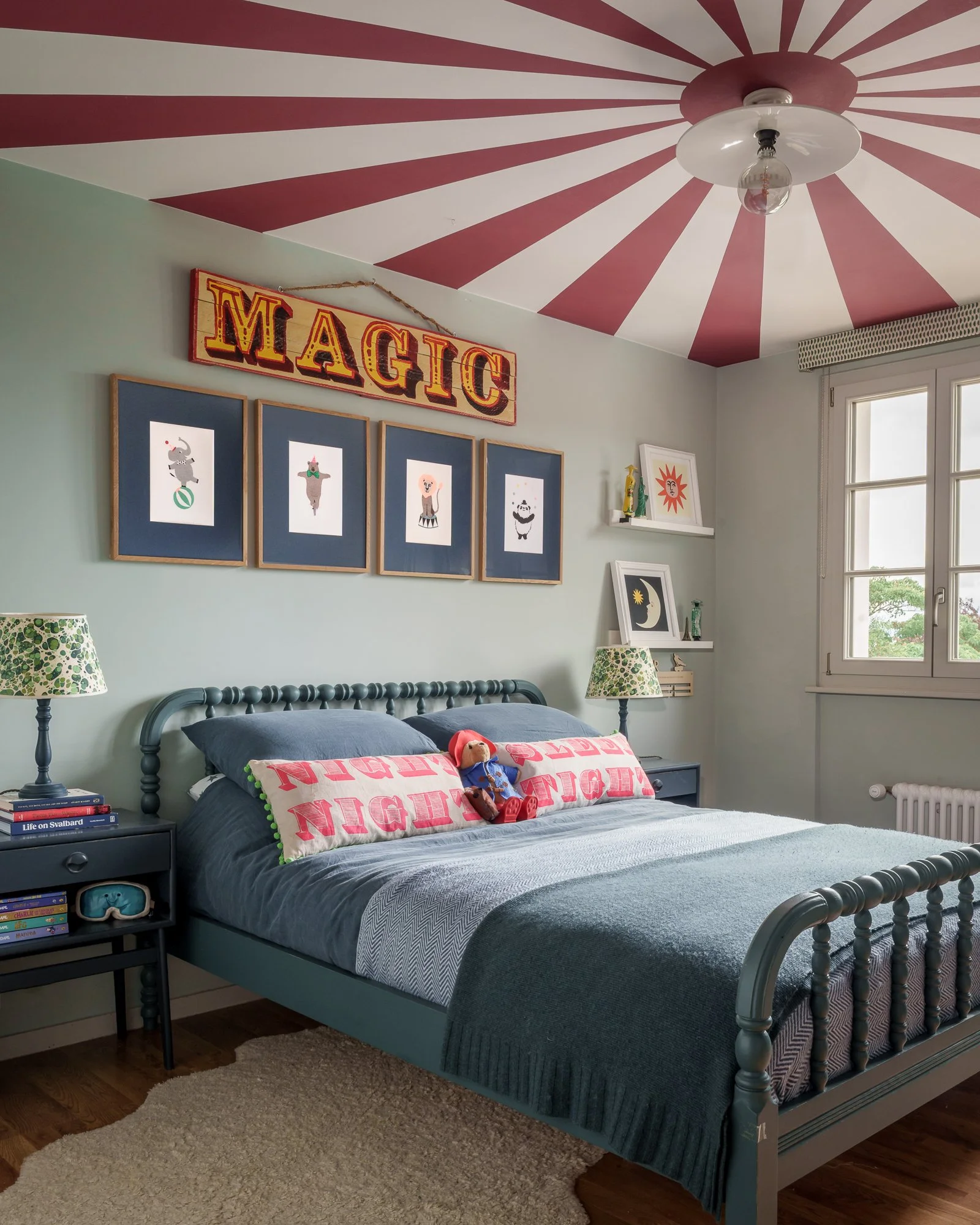 A bedroom with a blue metal bed, two pillows with pink and green designs, a teddy bear, nightstands with books and a mask, windows, and artwork including a sign that says 'MAGIC' with red and yellow letters and illustrations of circus animals. The ceiling has pink and white radial stripes with a light fixture. There are lamps on the nightstands and a window with a beige valance.