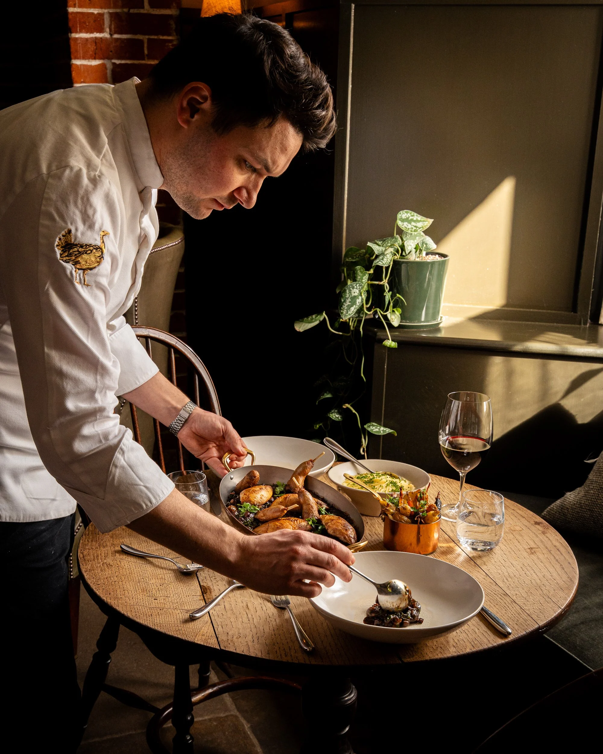 A chef serving a variety of gourmet dishes on a wooden table in a restaurant with natural light and a potted plant nearby.
