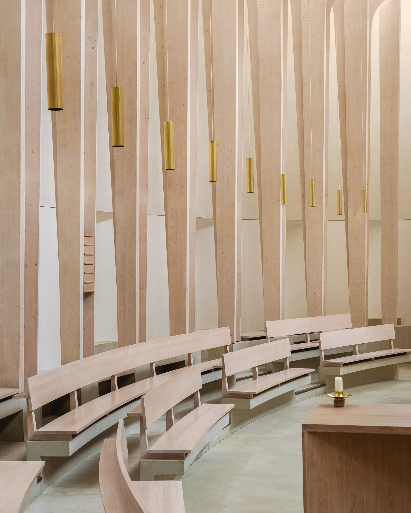 Interior of a religious or ceremonial space with curved wooden benches arranged in a semi-circle, a wooden wall with hanging cylindrical golden lights, a small table with a candle, and a wooden ladder against the wall.