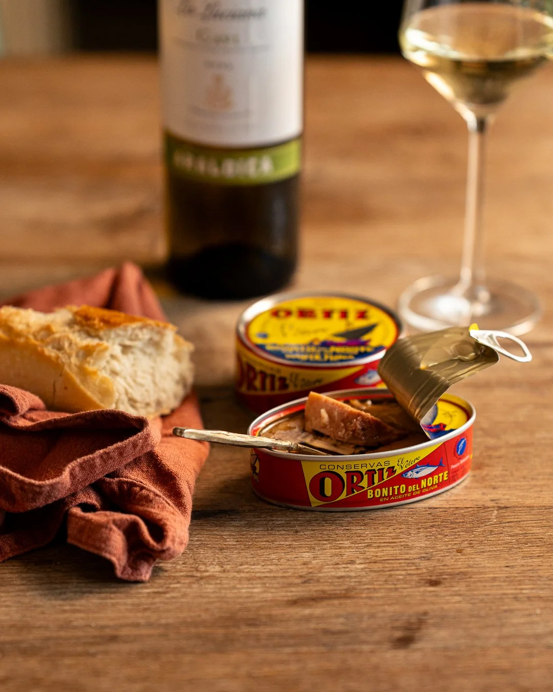 Open canned fish on a wooden table with bread, wine, and a glass of white wine in the background.