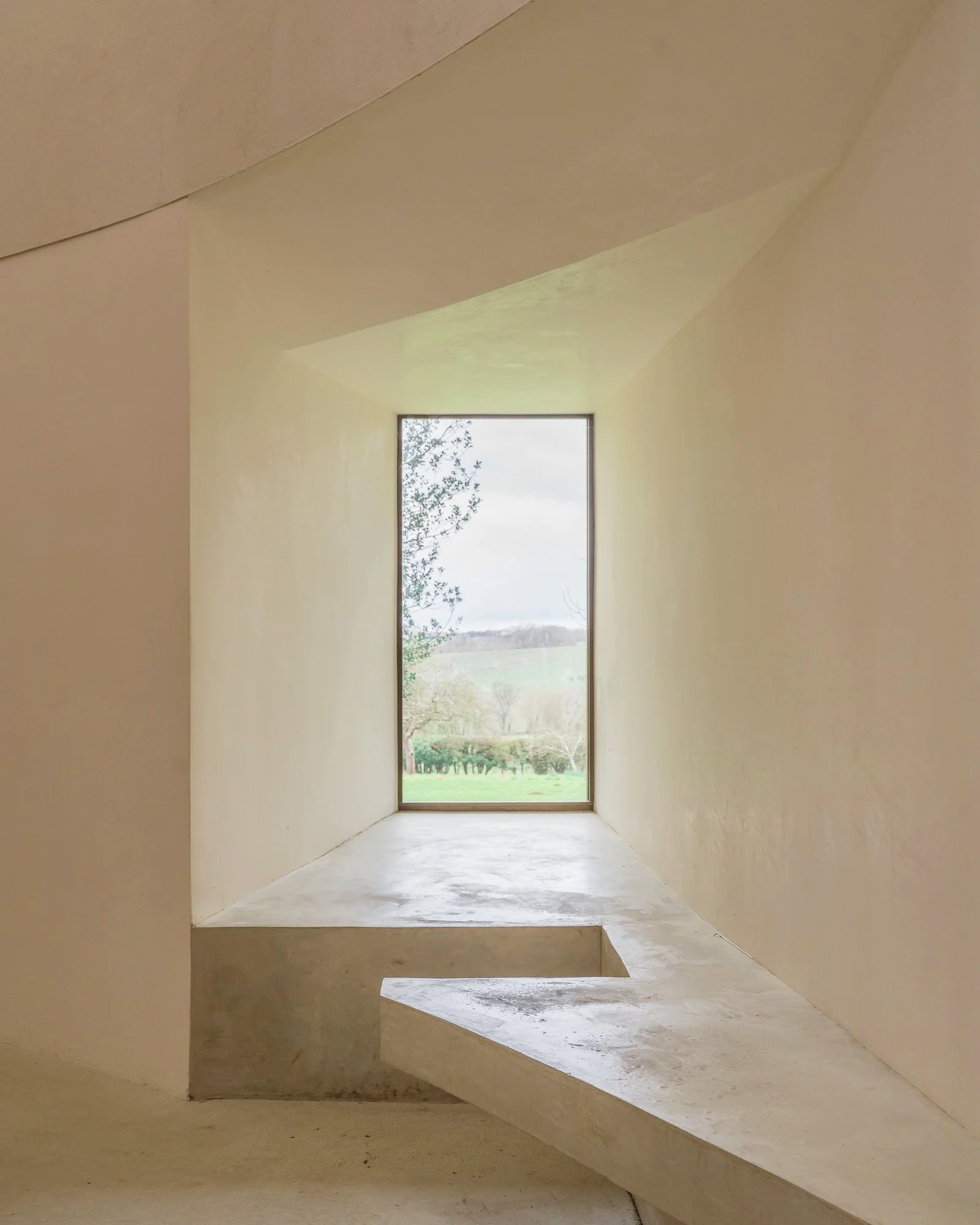 A modern building with a rectangular window revealing a view of trees and fields outside. The interior features smooth, light-colored walls and a concrete floor, with angular architectural details.