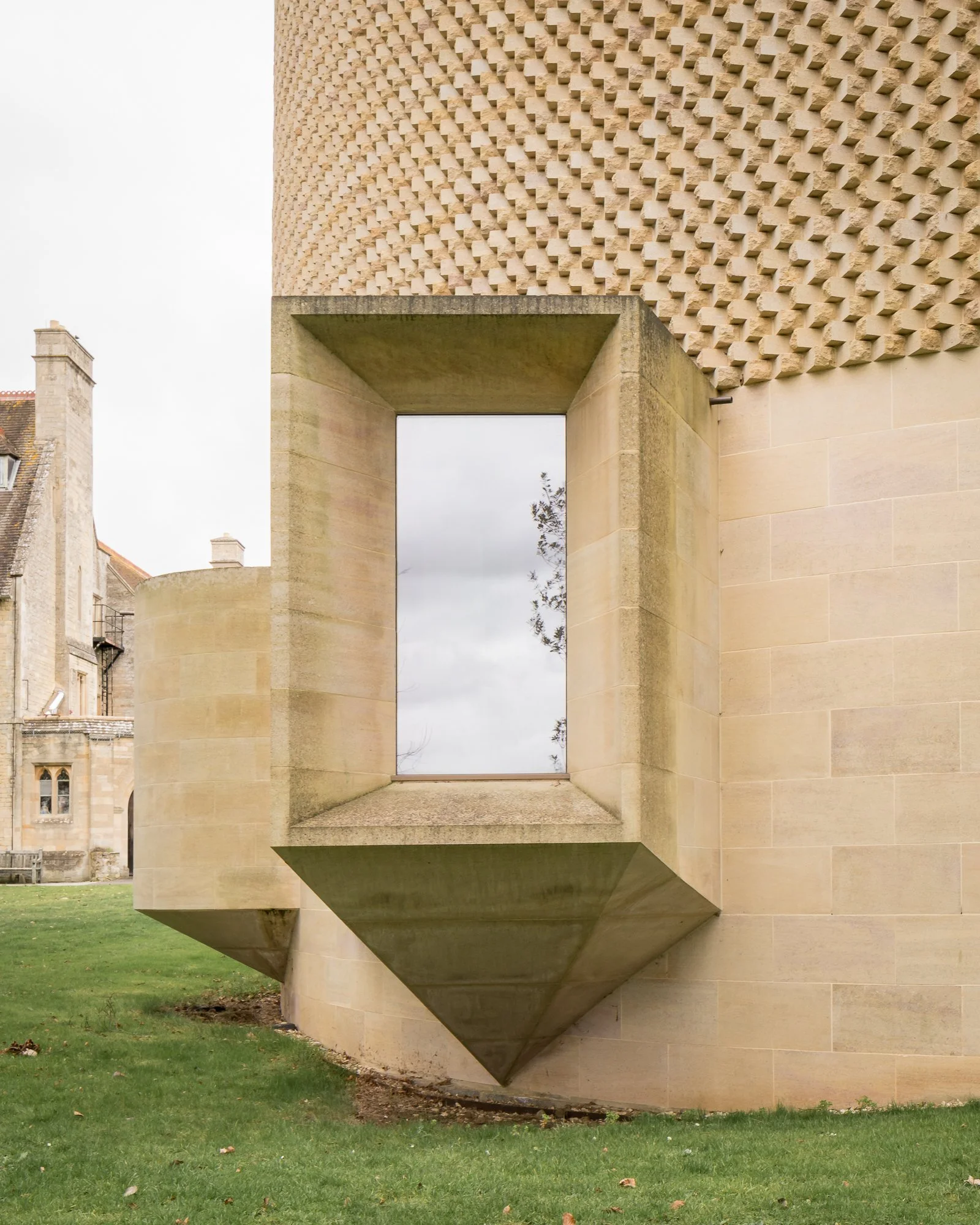 A modern architectural structure with a large, rectangular opening in the wall, made of beige stone. The building has a textured, patterned upper section with a grid of three-dimensional geometric shapes, and a smooth lower section. The structure is set on a grassy area with some fallen leaves, with a cloudy sky in the background.