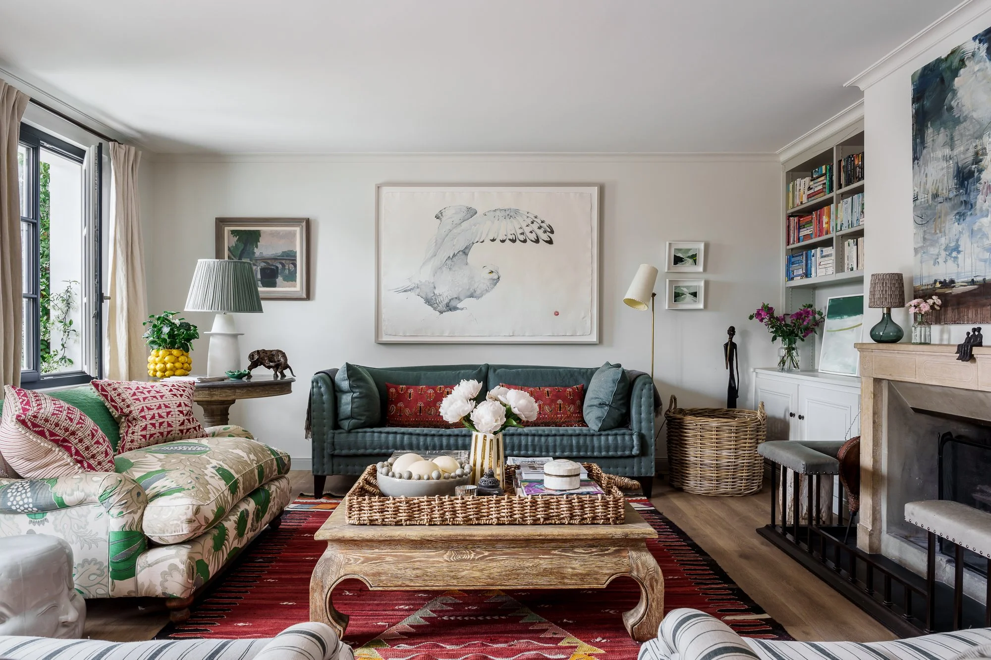 A living room with a floral-patterned couch, green velvet loveseat, and various decorative pillows. A wicker coffee table holds a vase of white flowers, and there are framed artworks, a bookshelf, and a fireplace with decorative items. Natural light comes in through a window with beige curtains.